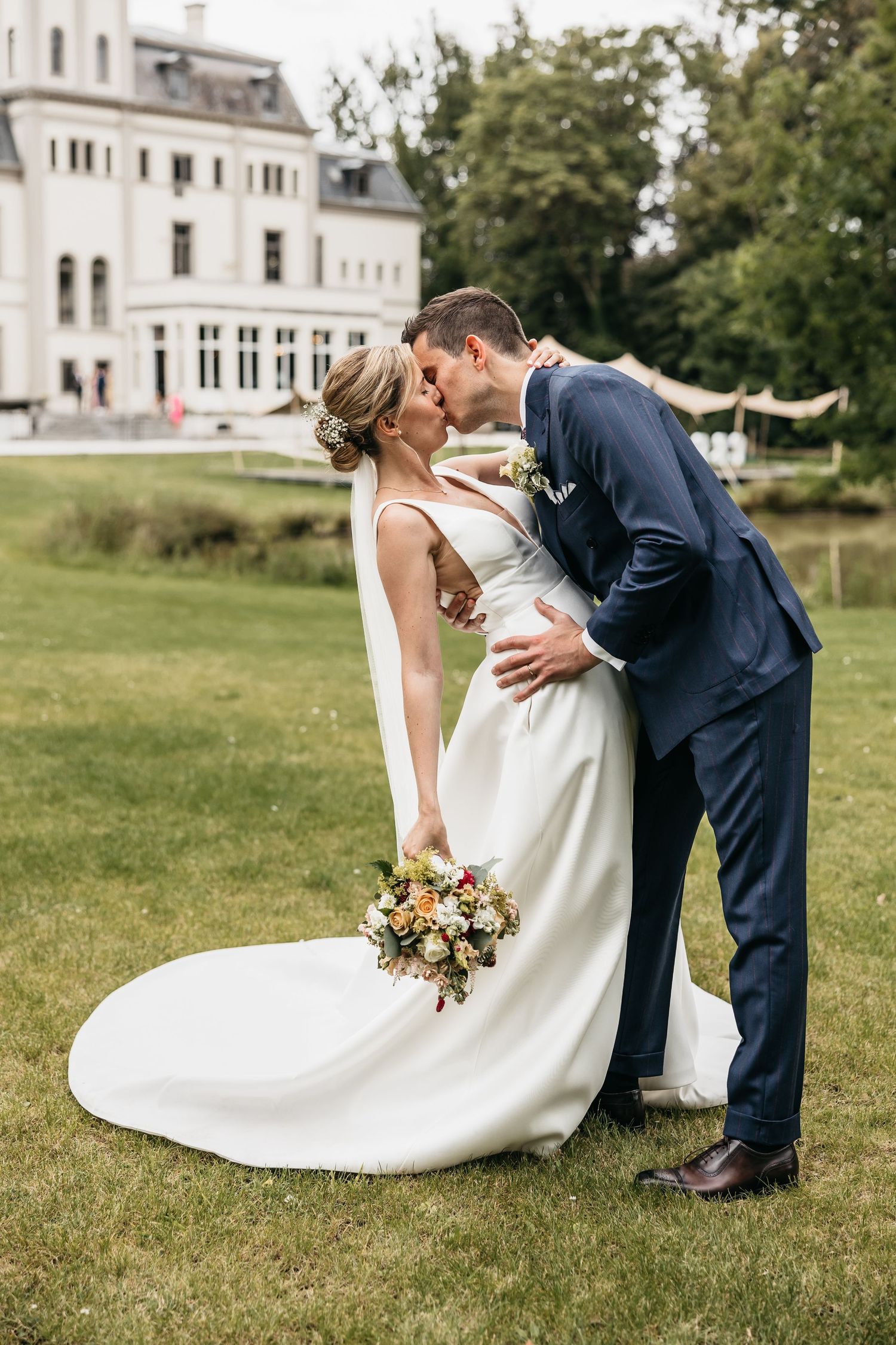 Bride and groom share a passionate kiss on green lawn with elegant white manor house behind.