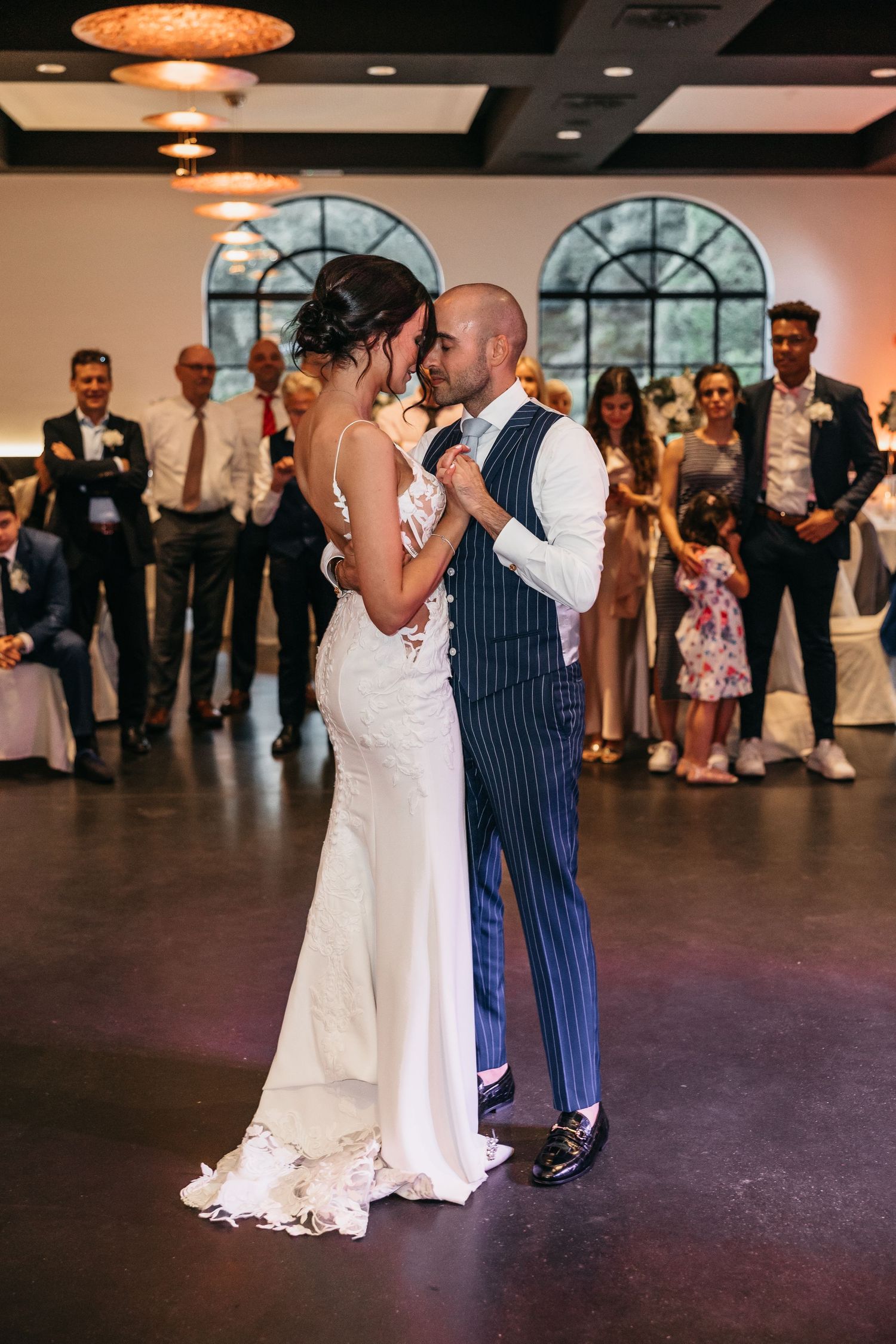 Bride in lace mermaid dress and groom in navy pinstripe suit dancing at wedding reception with guests watching.