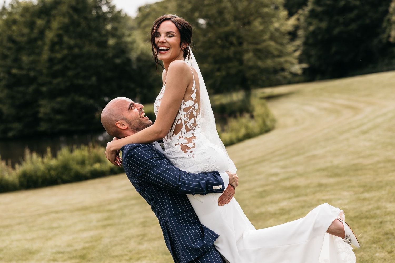 Joyful bride laughs as groom in navy pinstripe suit lifts her on lush green lawn.
