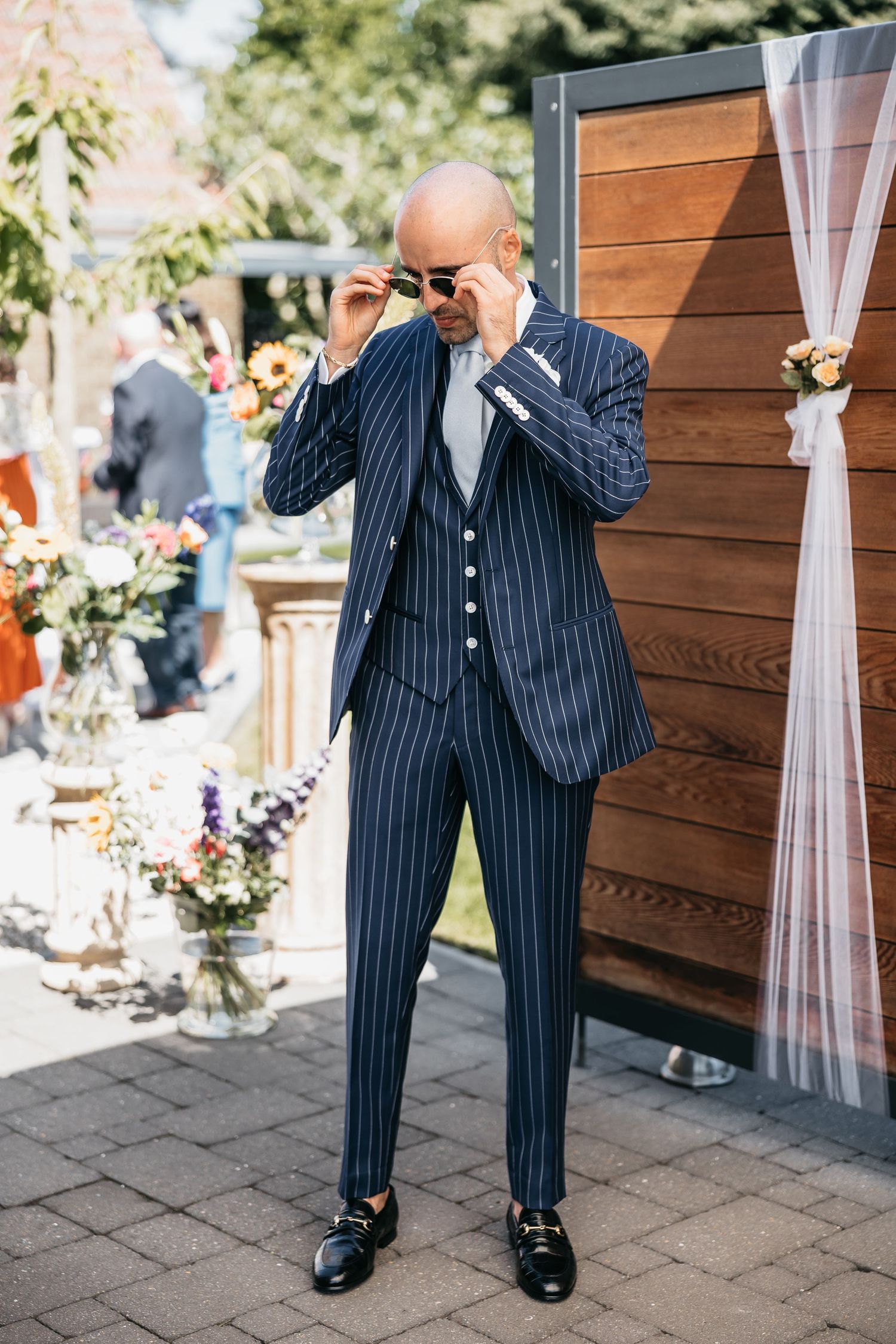 Bald groom adjusting sunglasses in navy pinstripe three-piece suit at outdoor wedding venue.