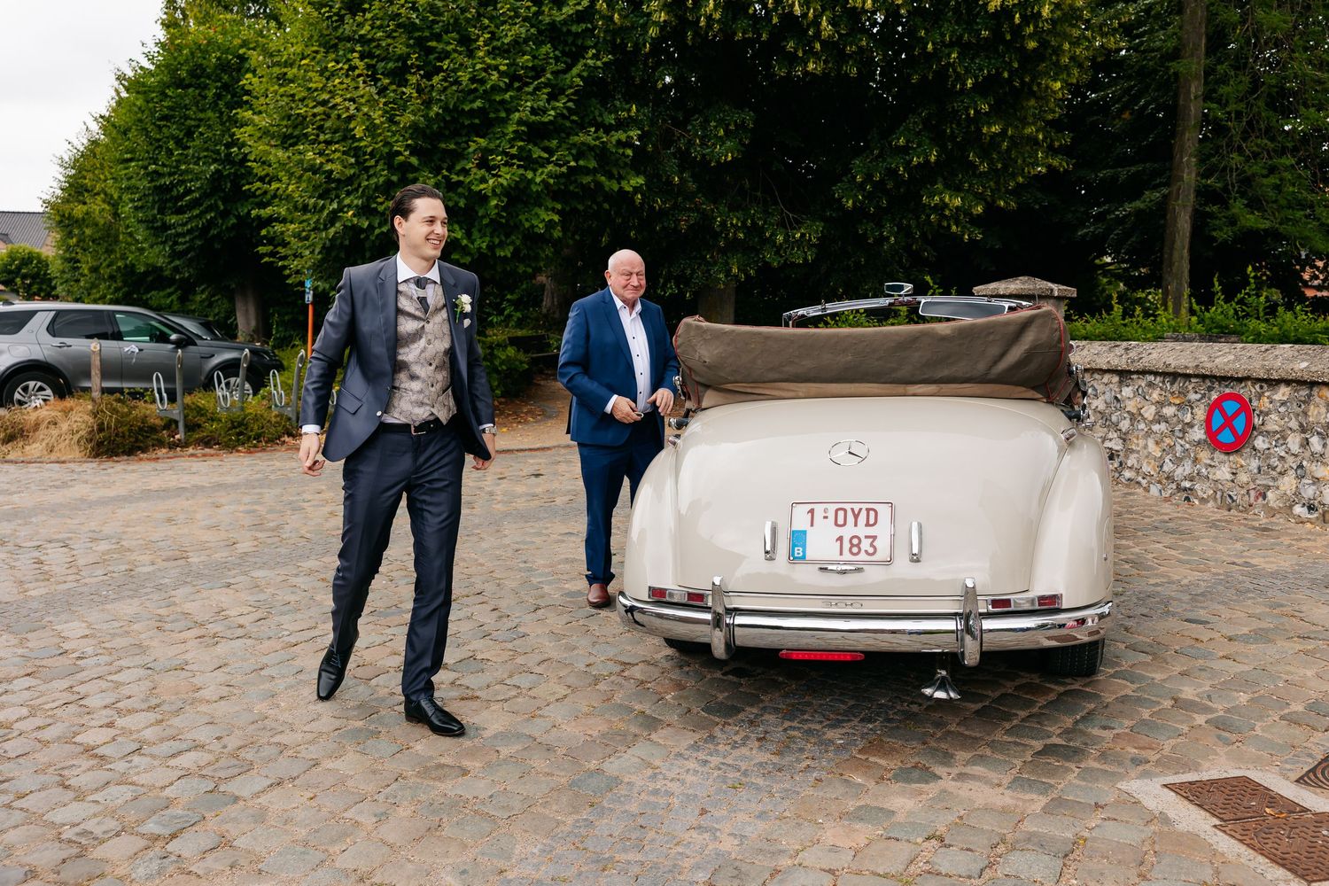 Groom in tan suit walks toward vintage cream Mercedes convertible with older gentleman.