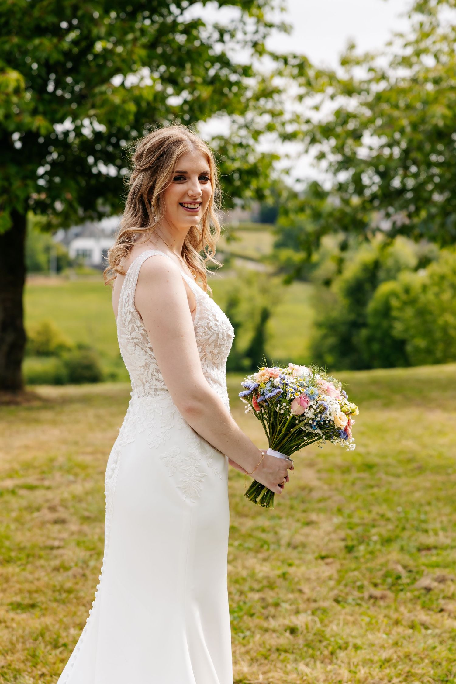 Smiling bride in sleeveless lace gown holding a wildflower bouquet, posing in a lush green outdoor setting.