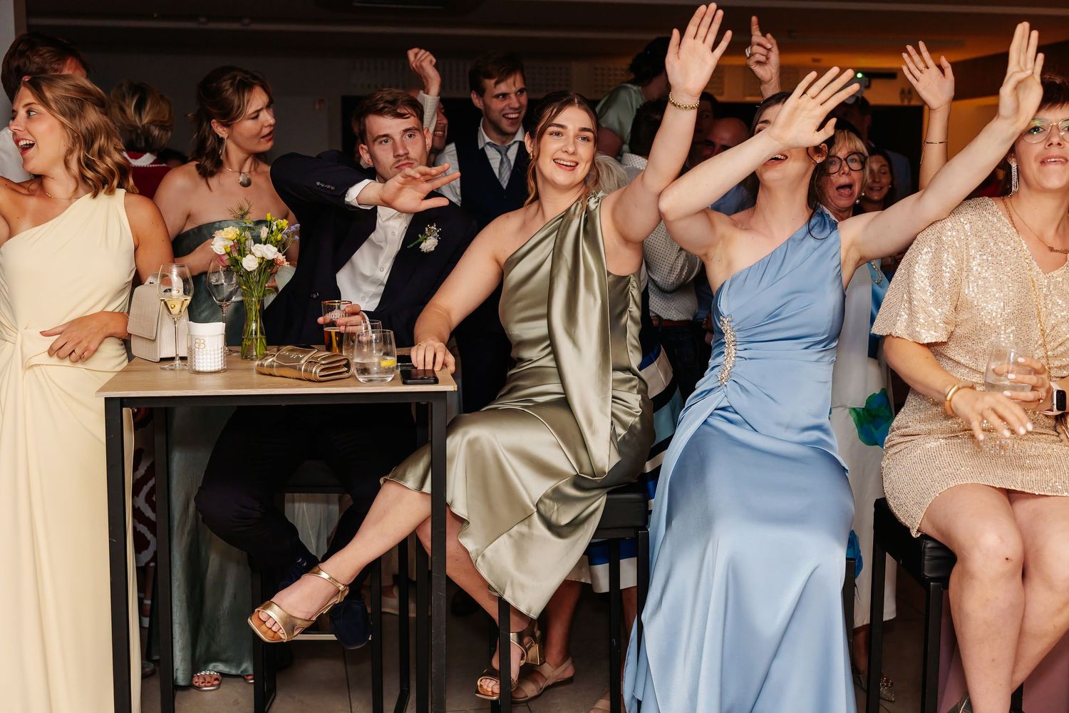 Smiling wedding guests in elegant dresses raise their hands during a fun reception celebration.