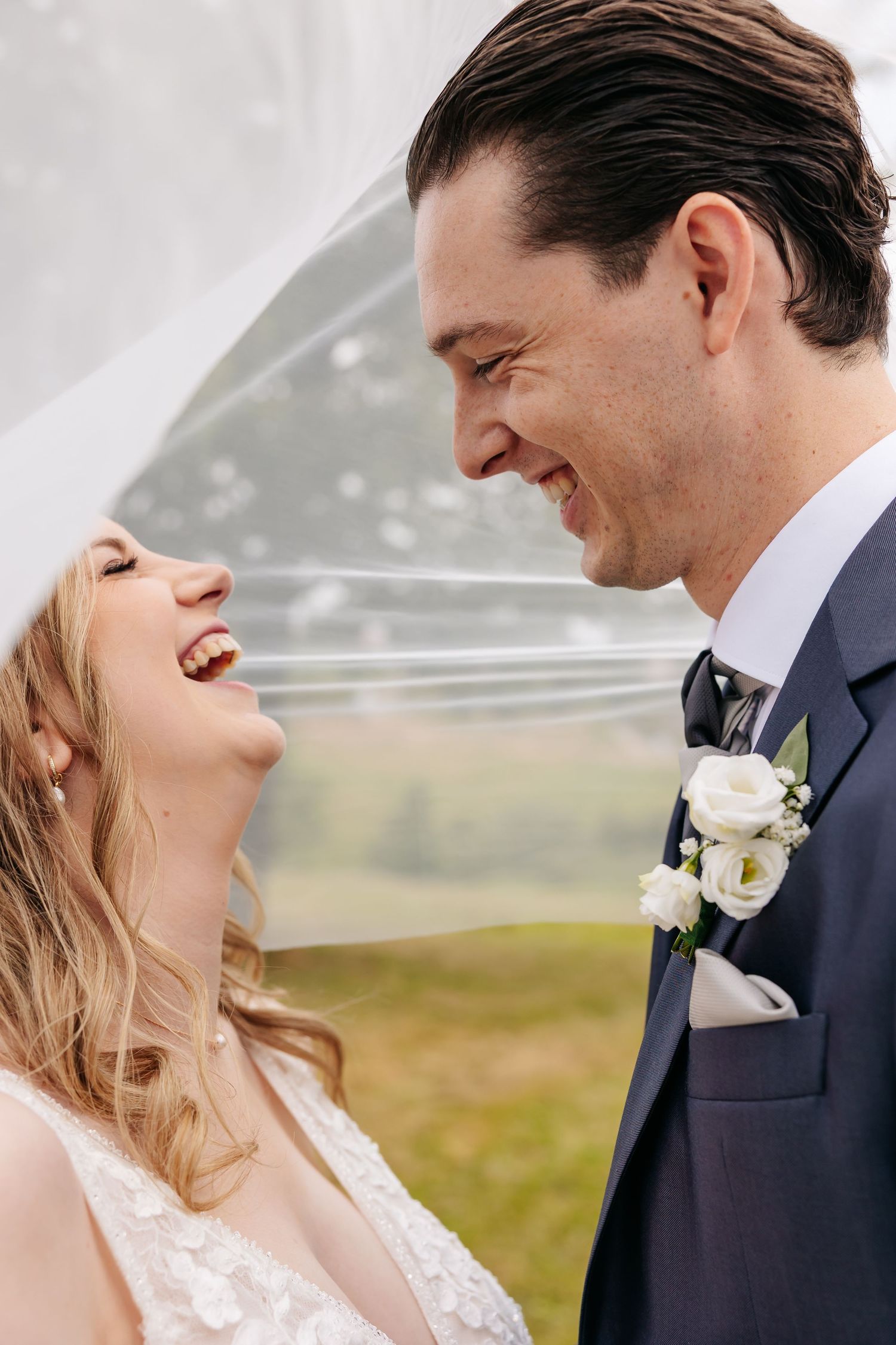 Joyful bride and groom laughing together under flowing wedding veil outdoors.