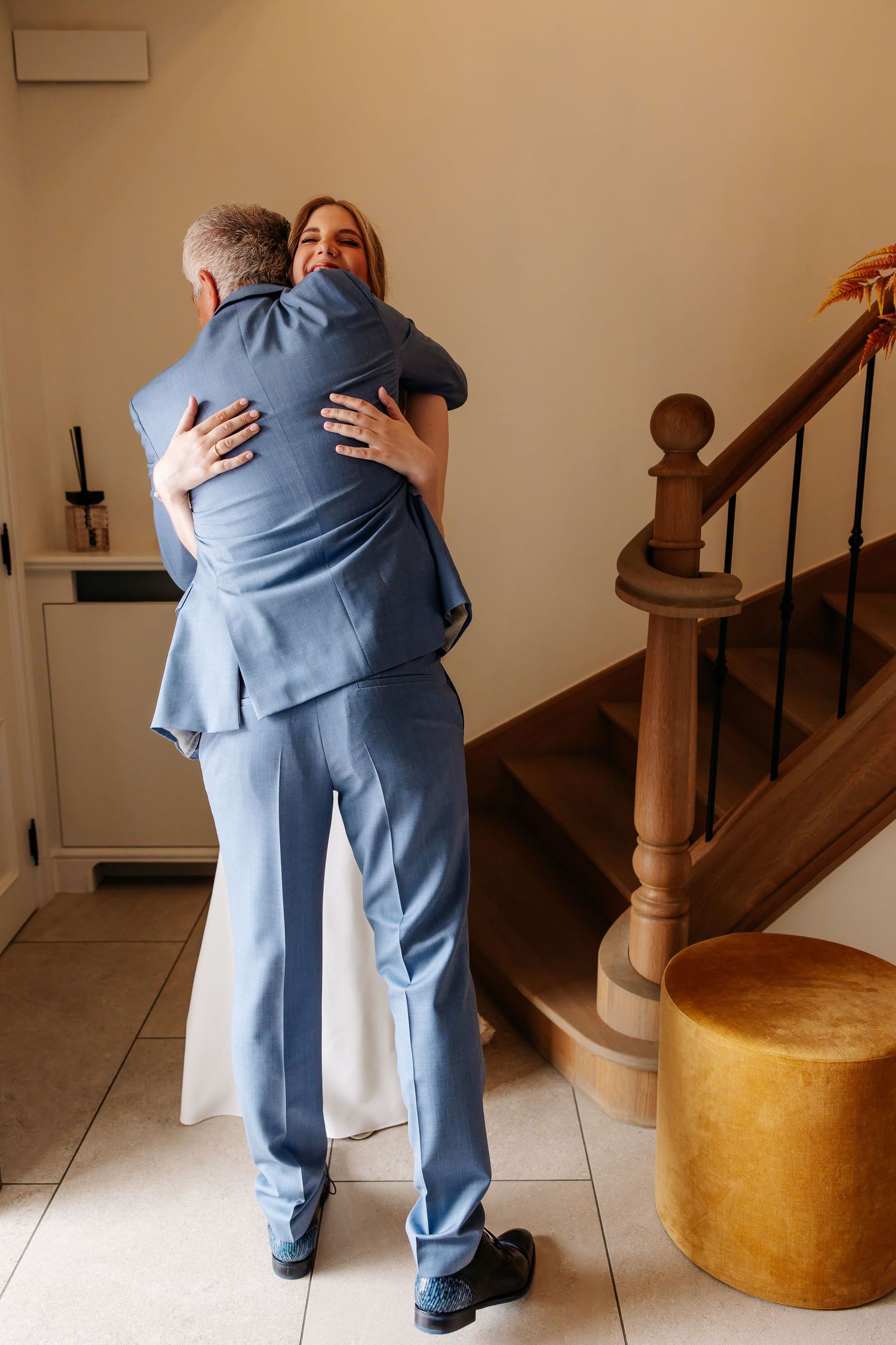 Bride in white gown embraces older man in blue suit near wooden staircase on wedding day.
