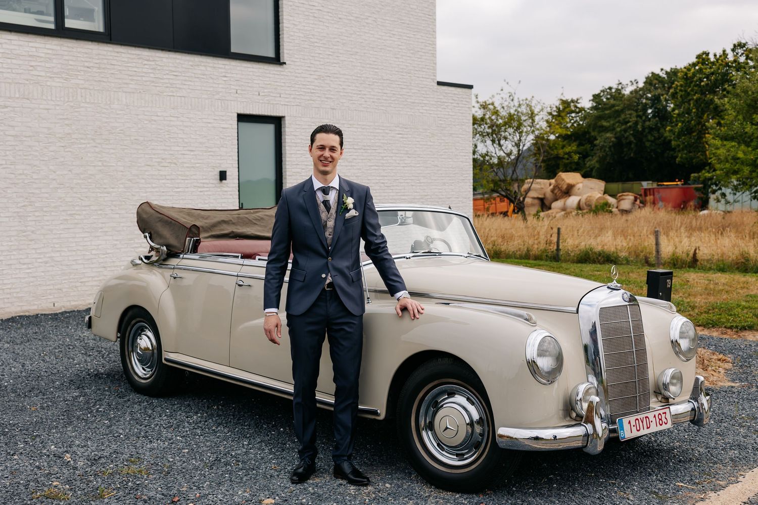 Groom in navy suit posing beside a vintage cream Mercedes convertible on a gravel driveway.