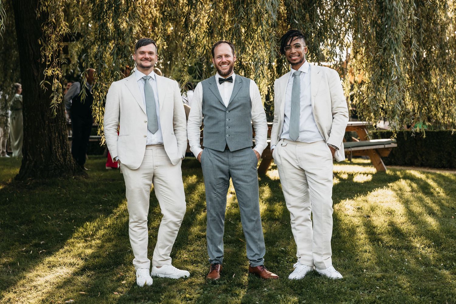 Groom in gray vest and bow tie stands with two groomsmen in cream suits under a willow tree.