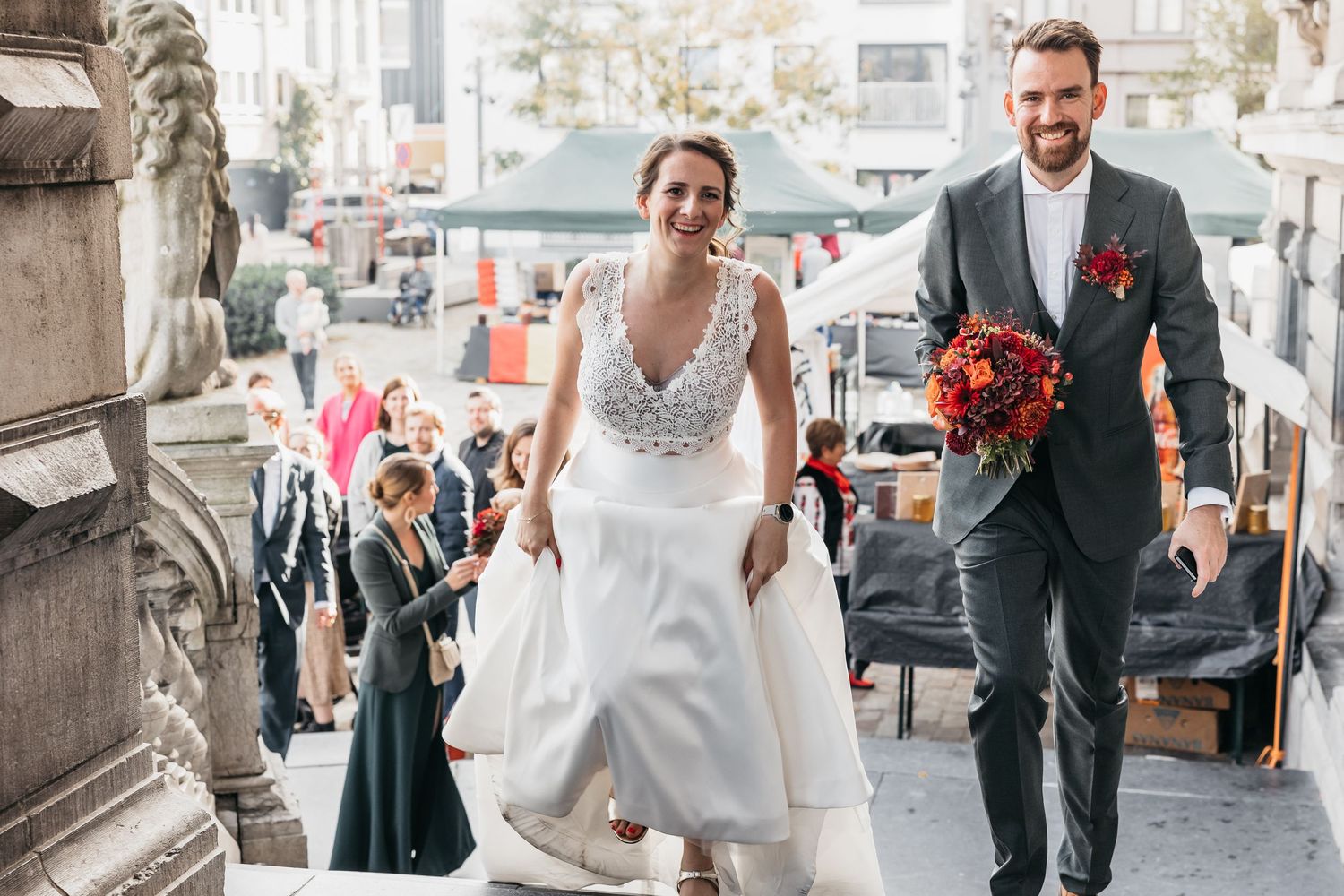 Bride and groom walk happily outside historic venue, groom carrying vibrant red and orange bouquet.