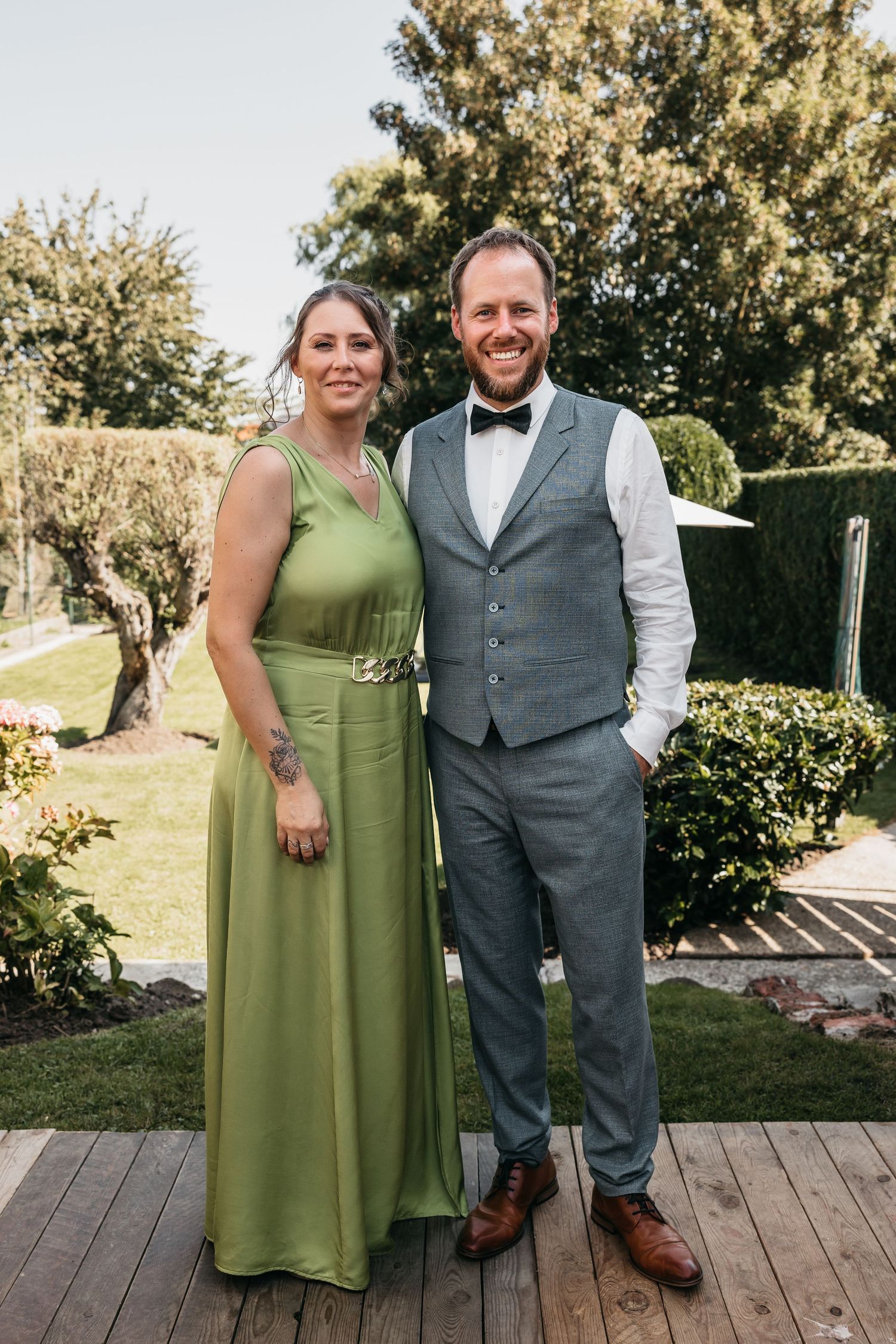 Couple posing at a wedding, woman in elegant green gown and man in gray suit with bow tie on a wooden deck.