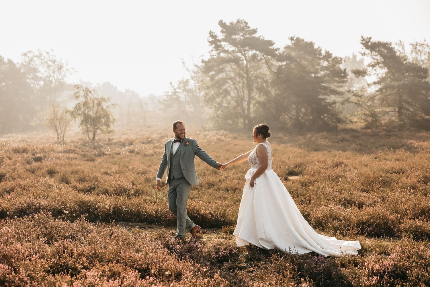 Bride in flowing white gown and groom in grey suit walk hand in hand through a golden misty heathland.