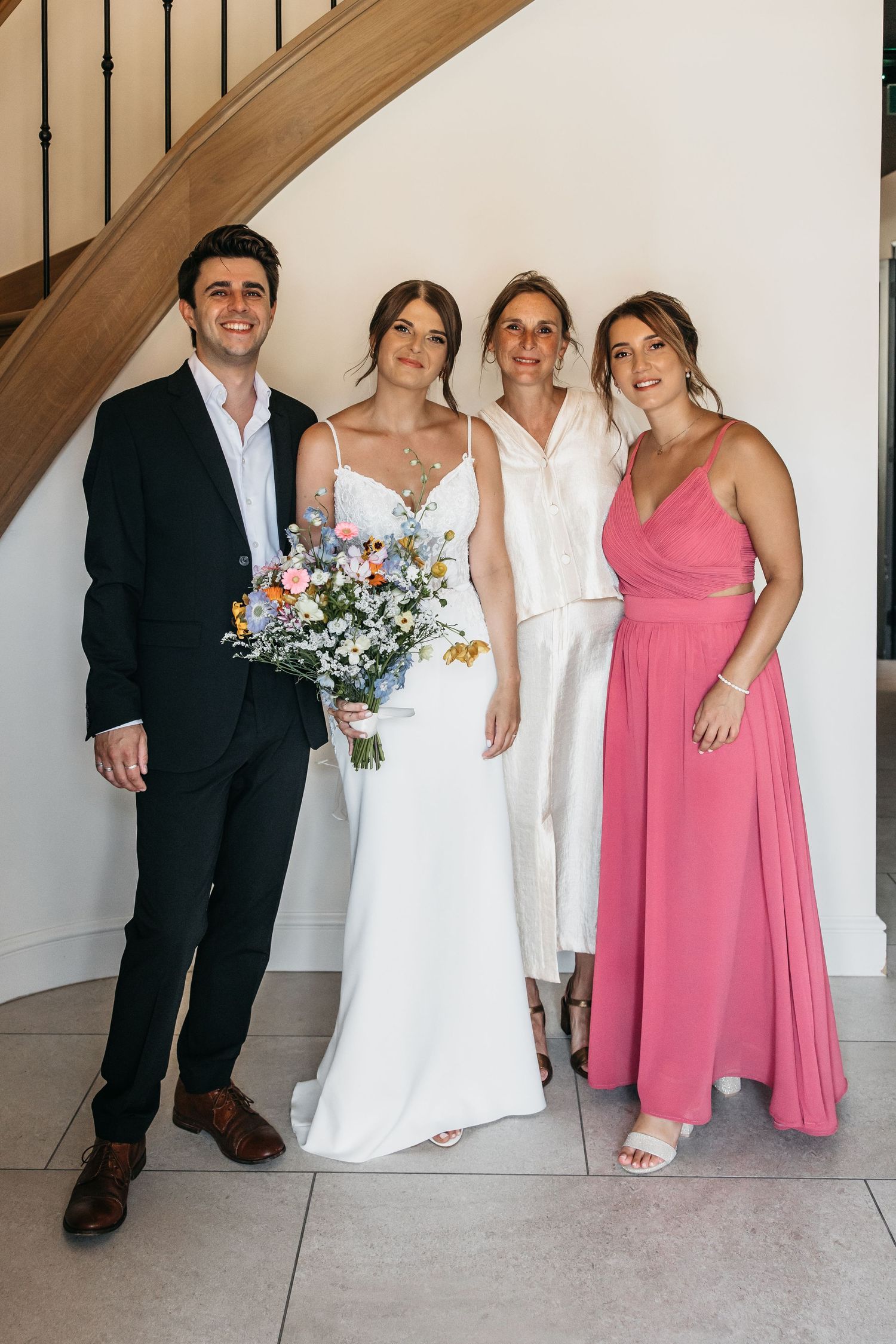 Bride with wildflower bouquet poses with three smiling family members beside curved wooden staircase.