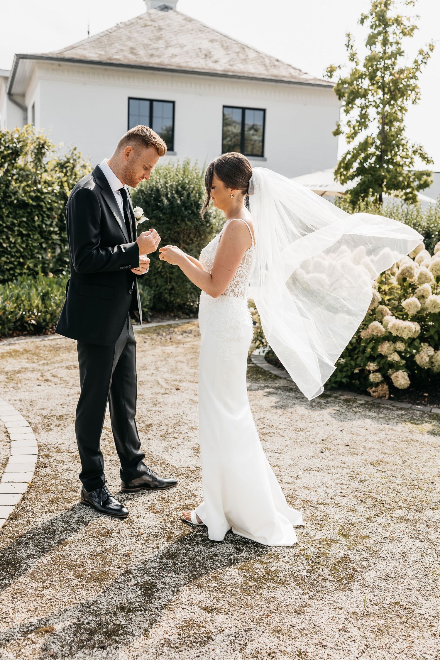 Bride and groom exchange rings outdoors near white building with hydrangeas, bride's veil flowing in the breeze.