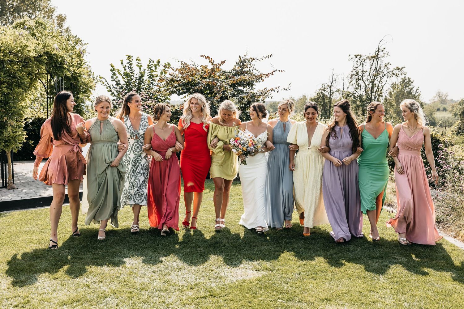 Bride and bridesmaids in colorful mismatched dresses laughing together on green lawn at outdoor wedding.