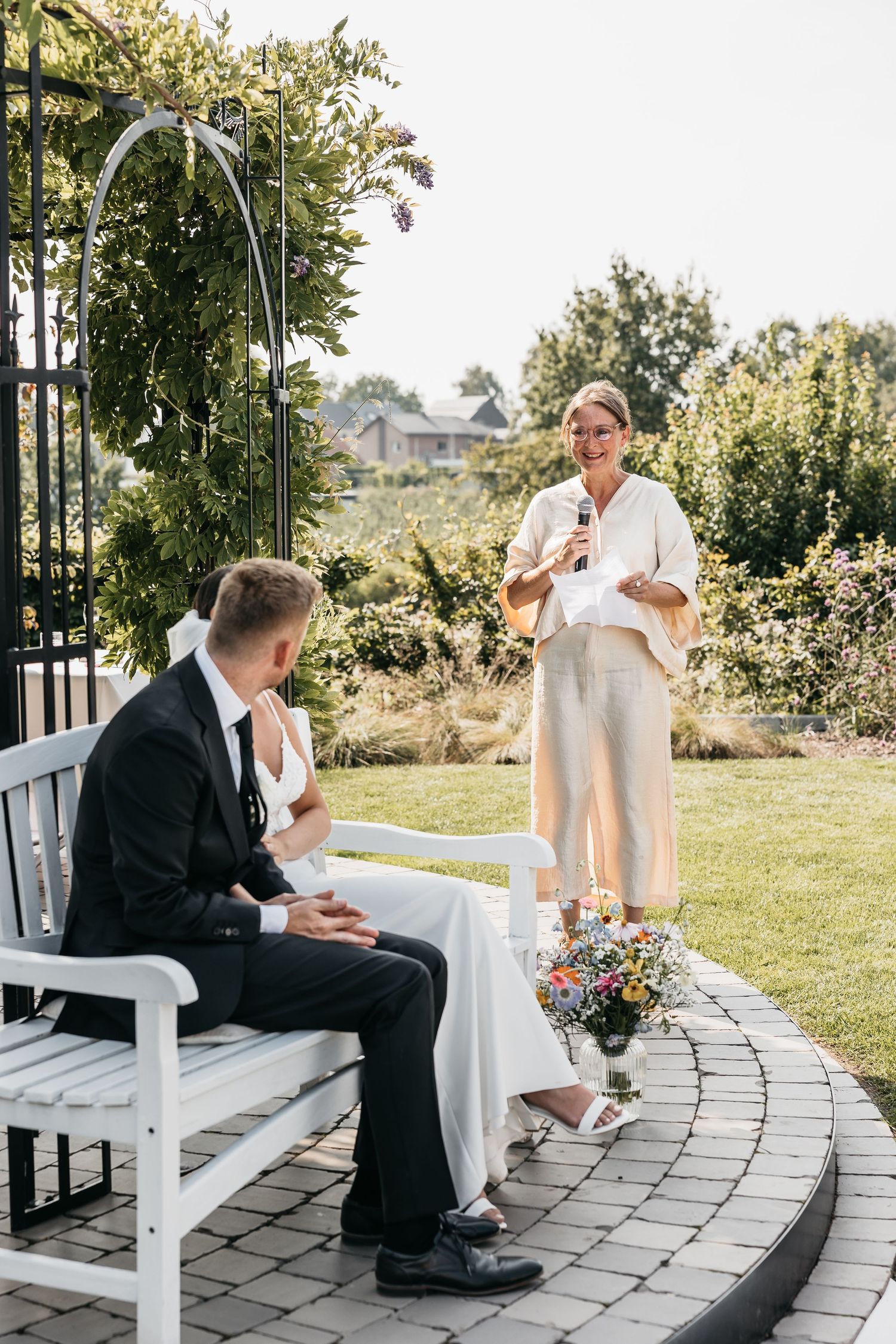 Wedding officiant in white suit speaking into microphone during outdoor garden ceremony with couple.