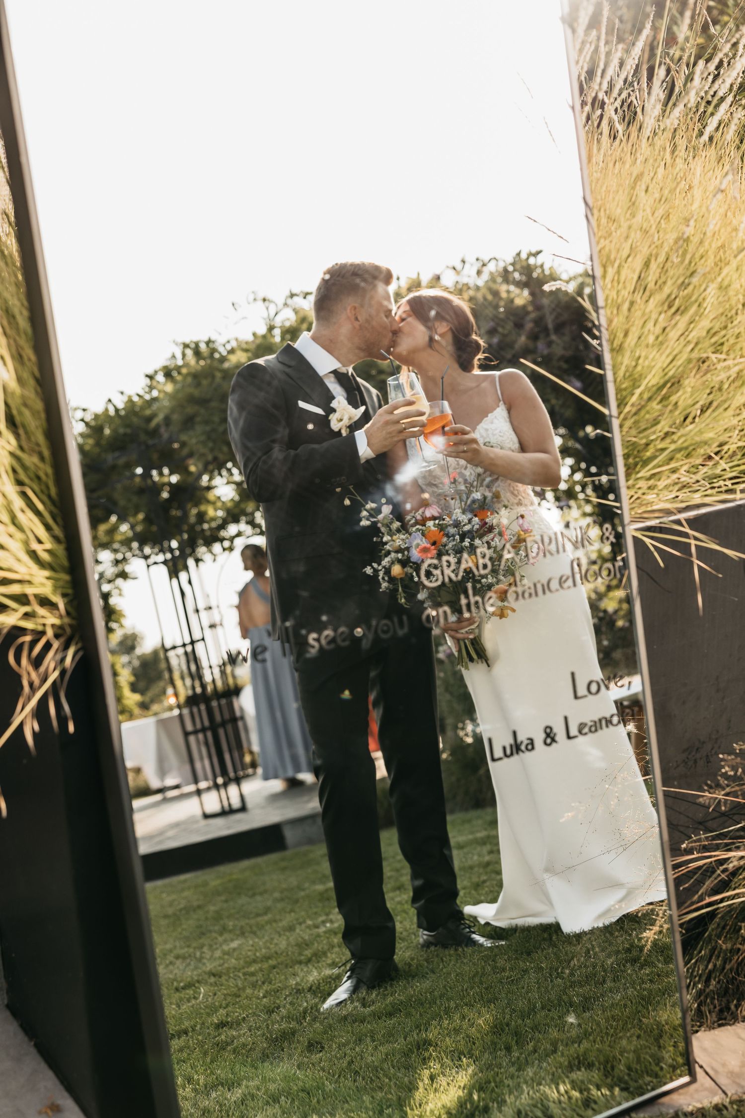 Bride and groom share a kiss while holding champagne glasses at an outdoor wedding ceremony.