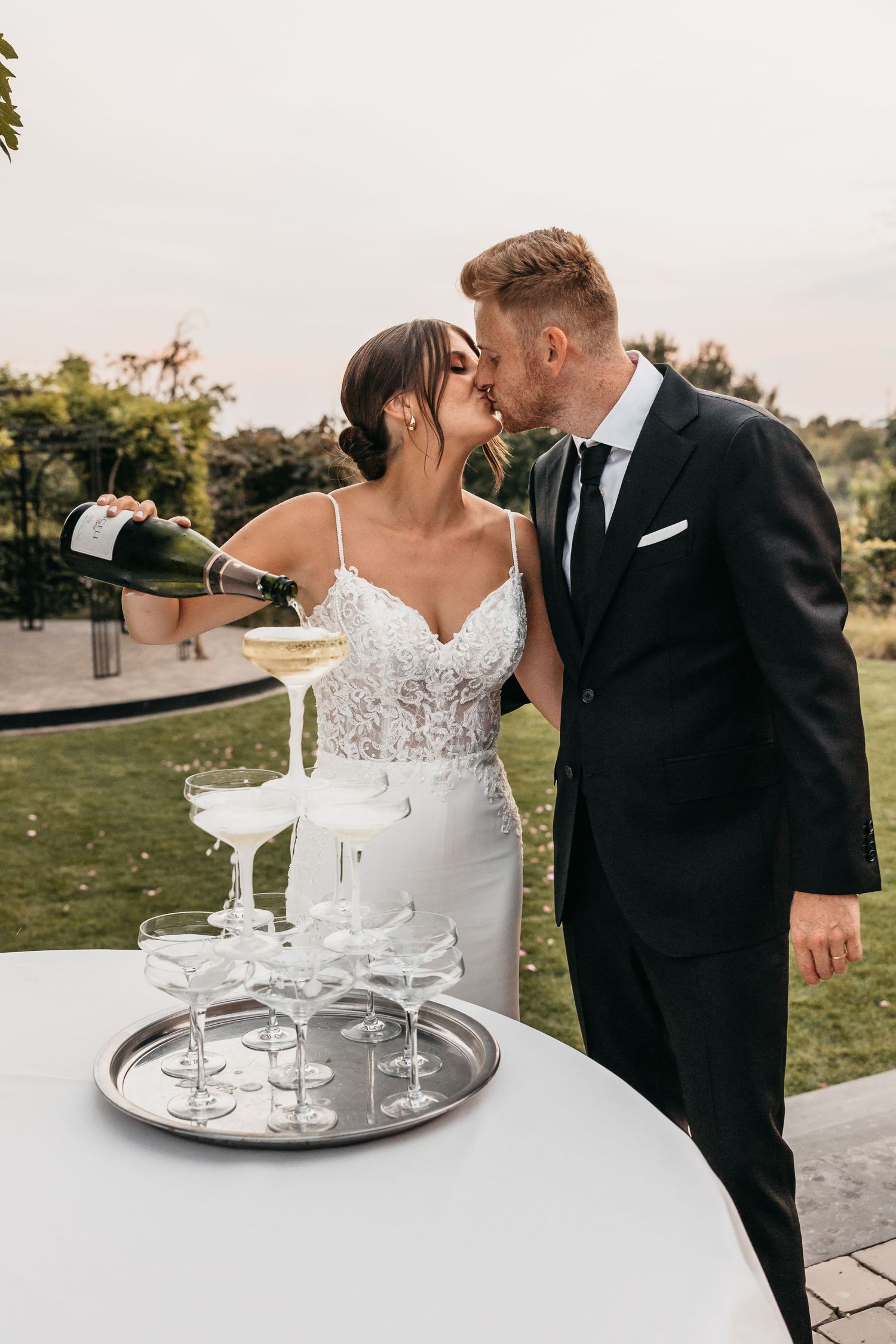 Bride and groom kissing while pouring champagne tower at outdoor wedding reception.
