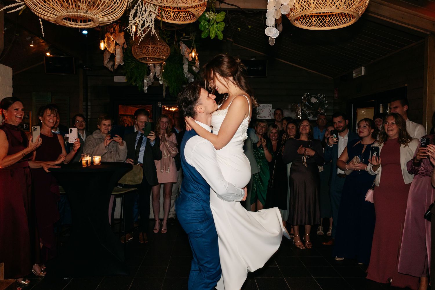 Groom lifting bride on dance floor surrounded by wedding guests during first dance celebration.
