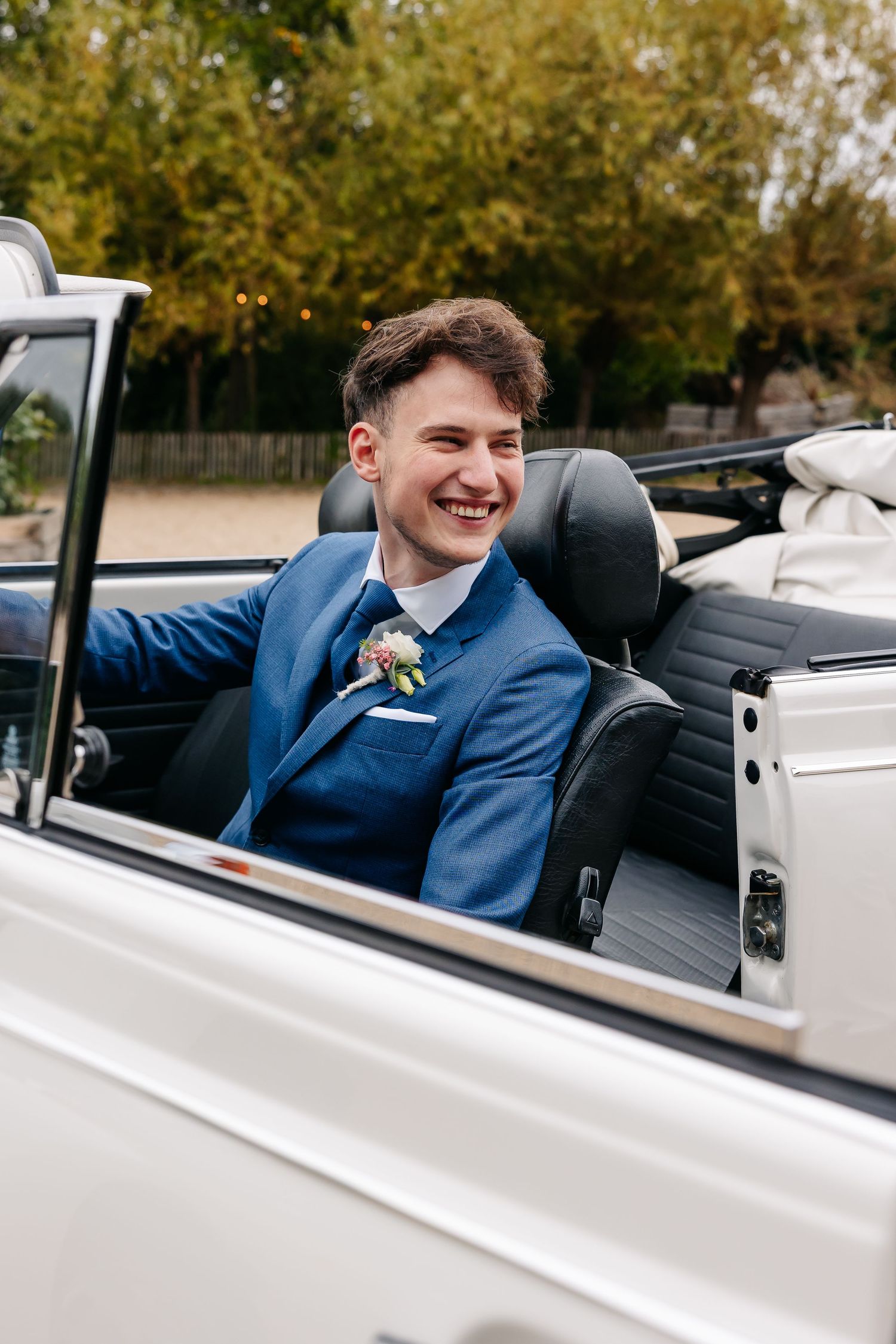 Smiling groom in blue suit with boutonniere seated in white vintage convertible wedding car.