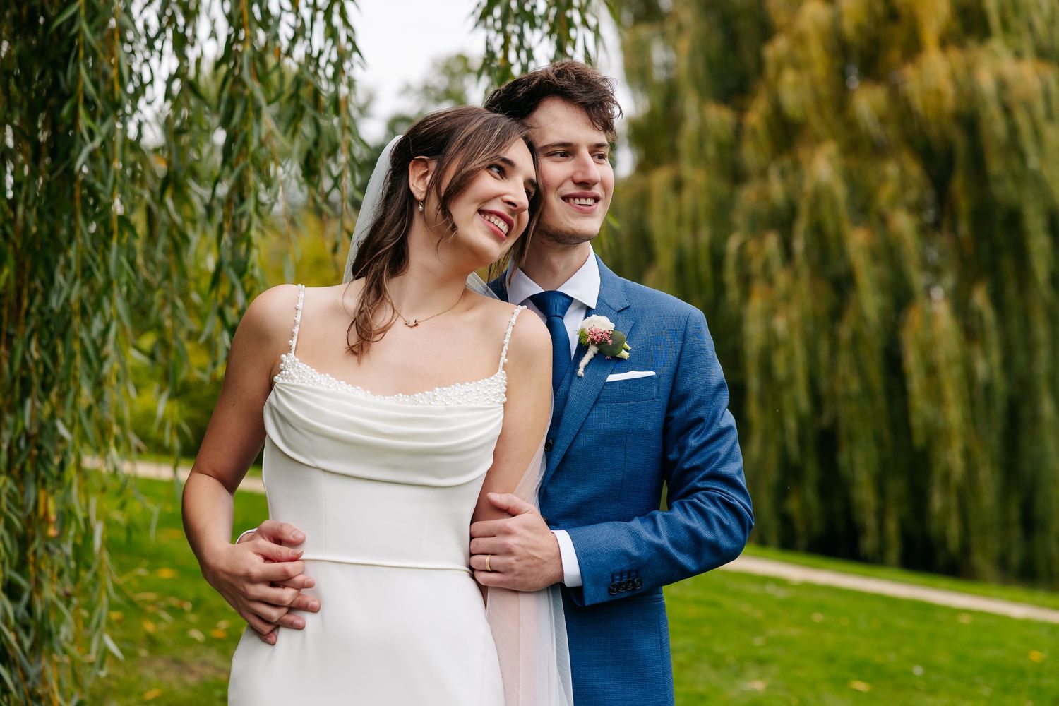 Smiling bride and groom in blue suit share joyful moment together under weeping willow tree.