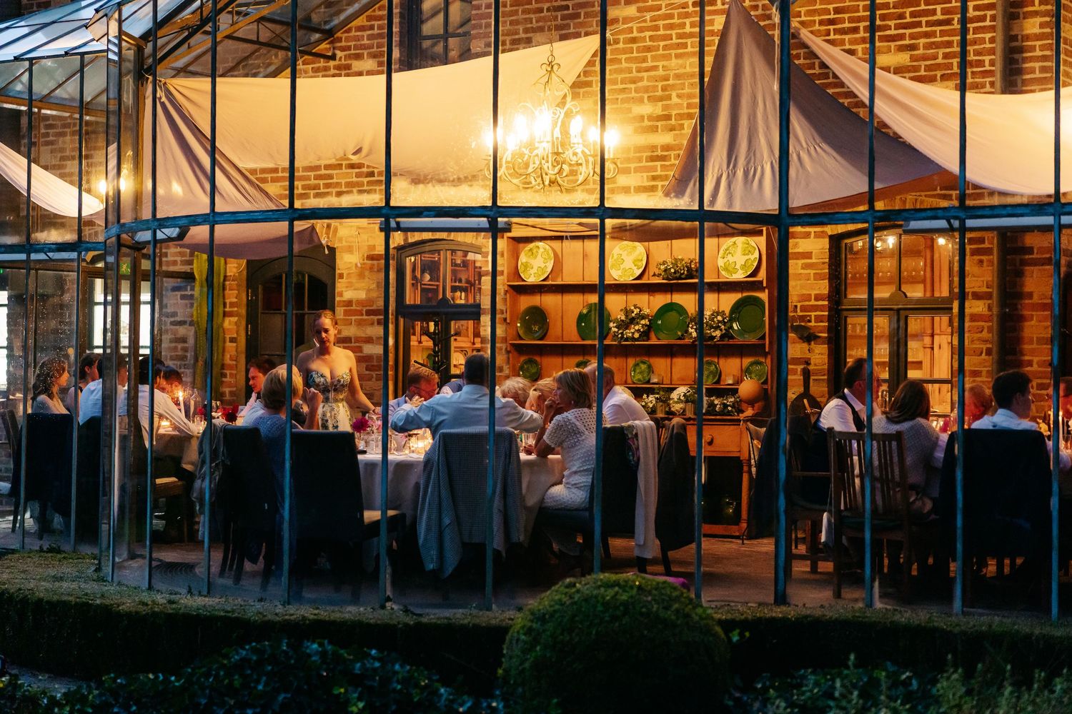 Wedding guests dining elegantly inside a glass conservatory glowing warmly at dusk.