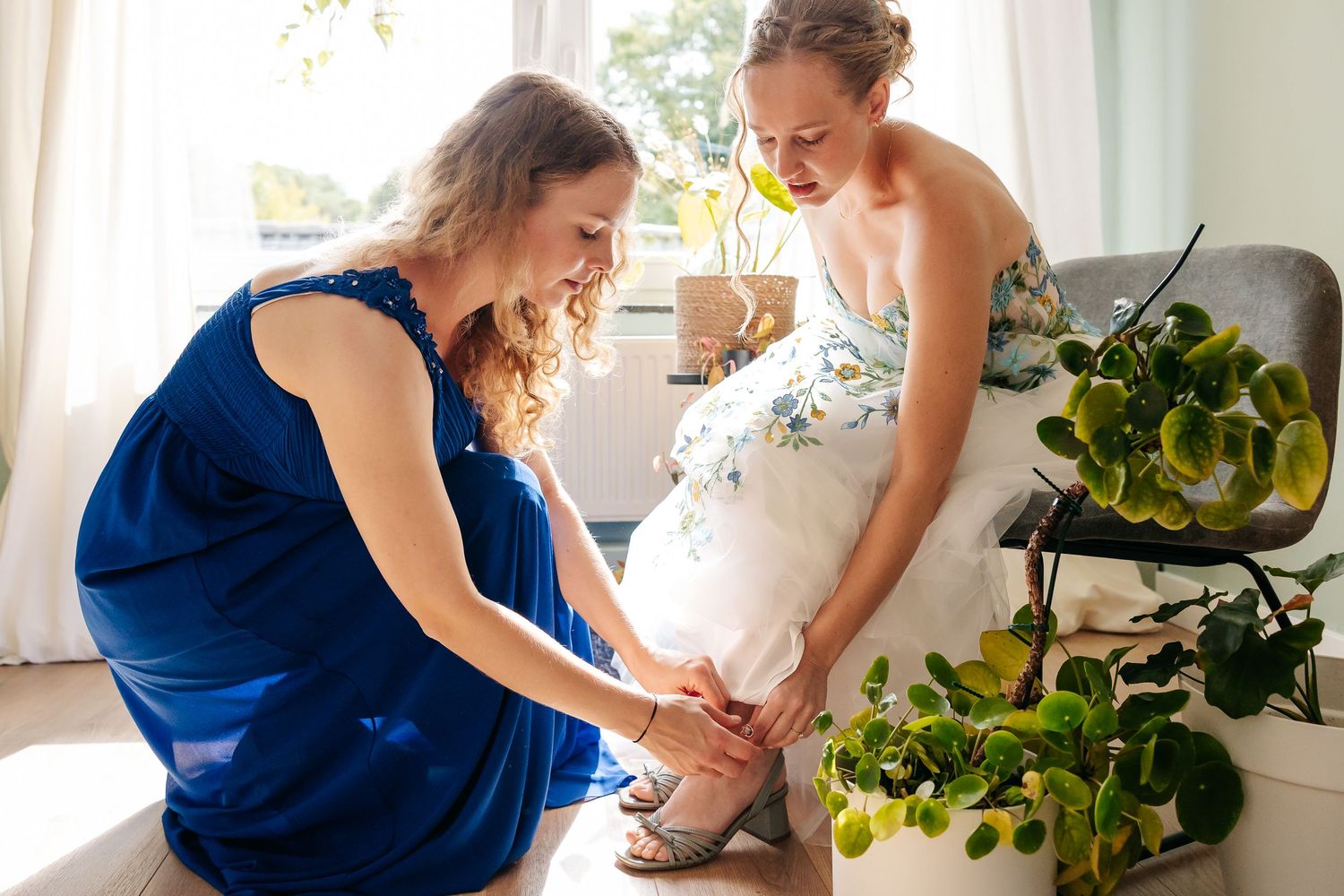 Bridesmaid in blue dress kneeling to help bride put on wedding shoes in bright room.