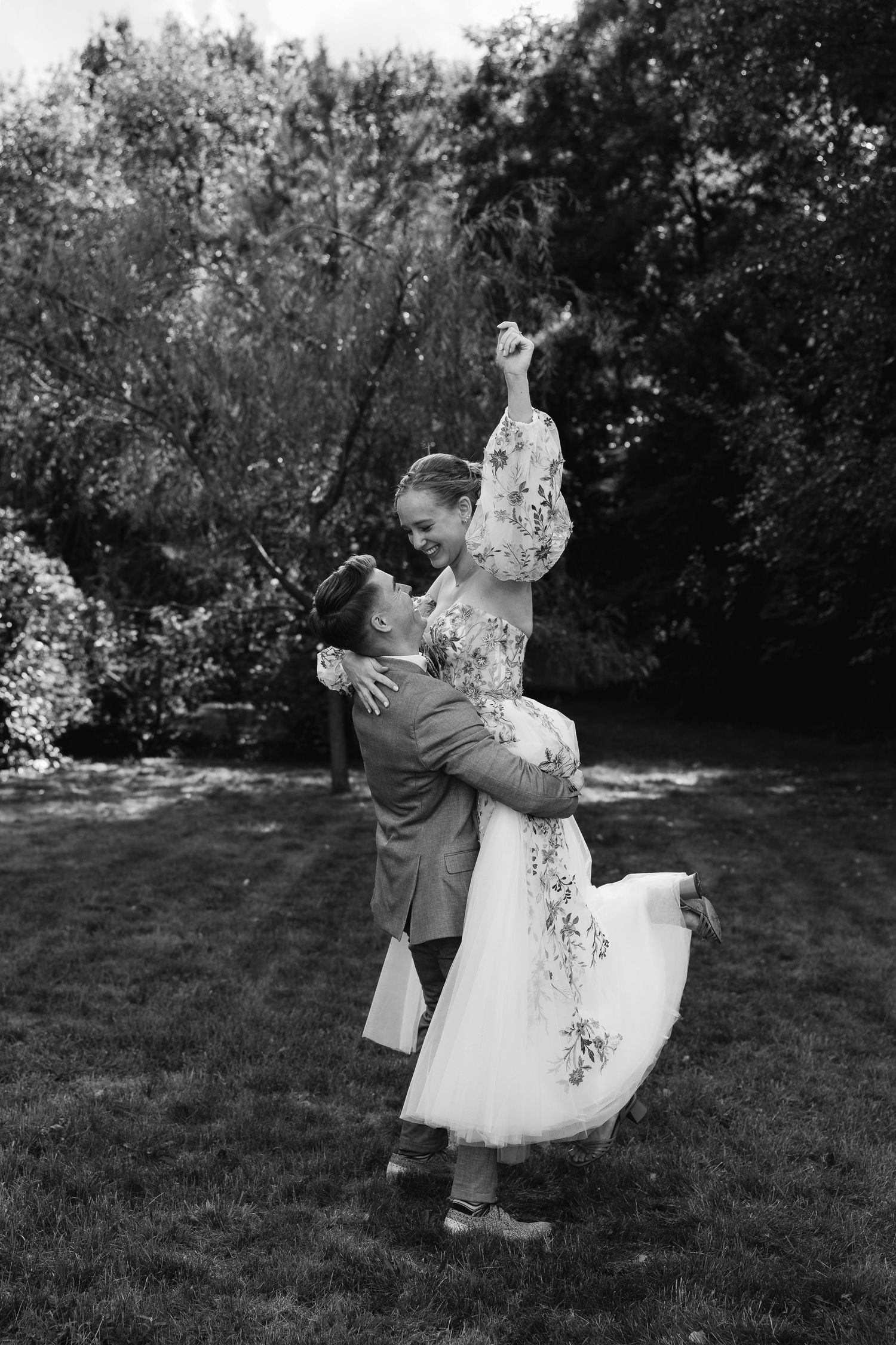 Black and white photo of groom lifting bride in floral dress in a romantic garden wedding moment.