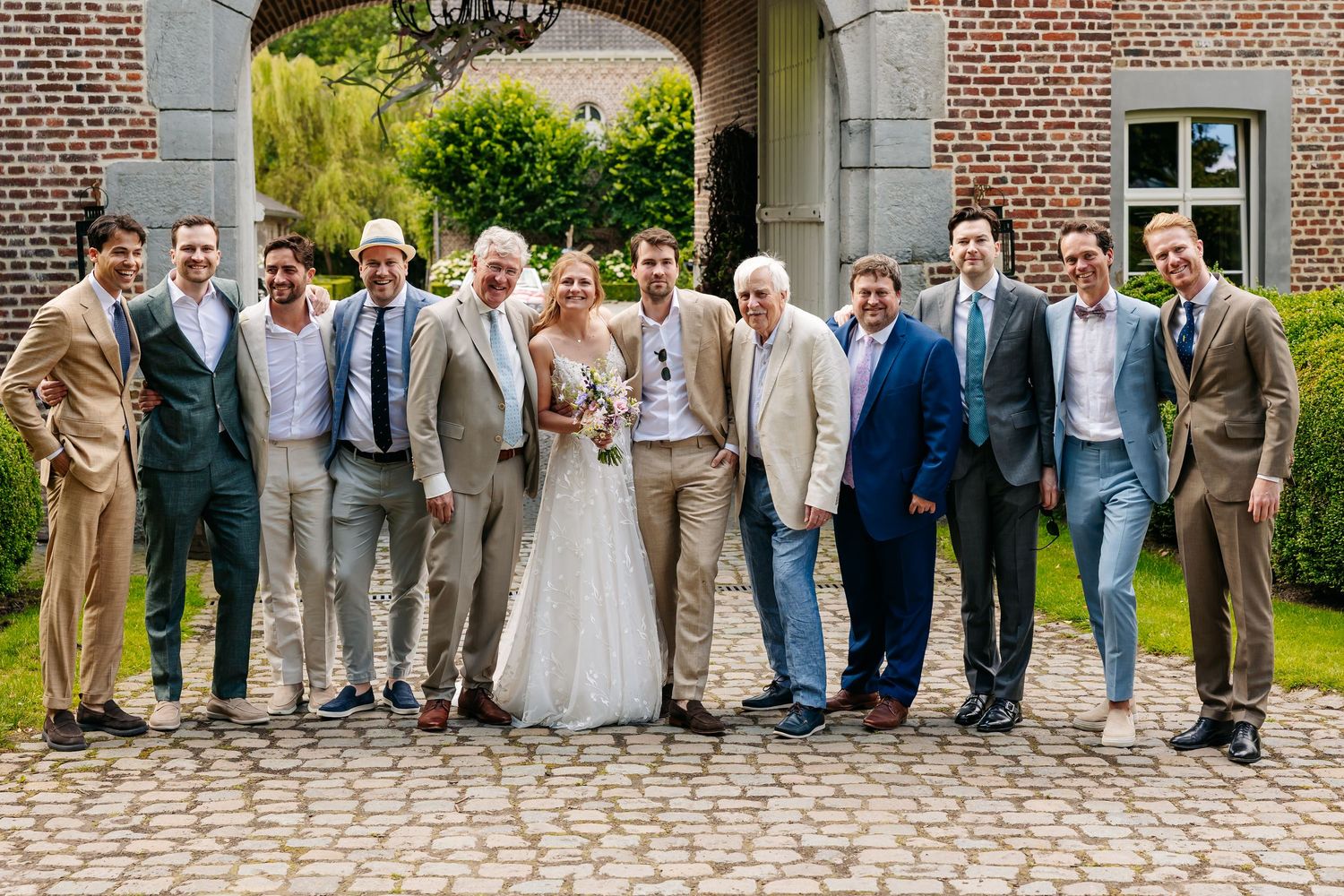 Bride posing with large group of men in suits at outdoor wedding on cobblestone courtyard.
