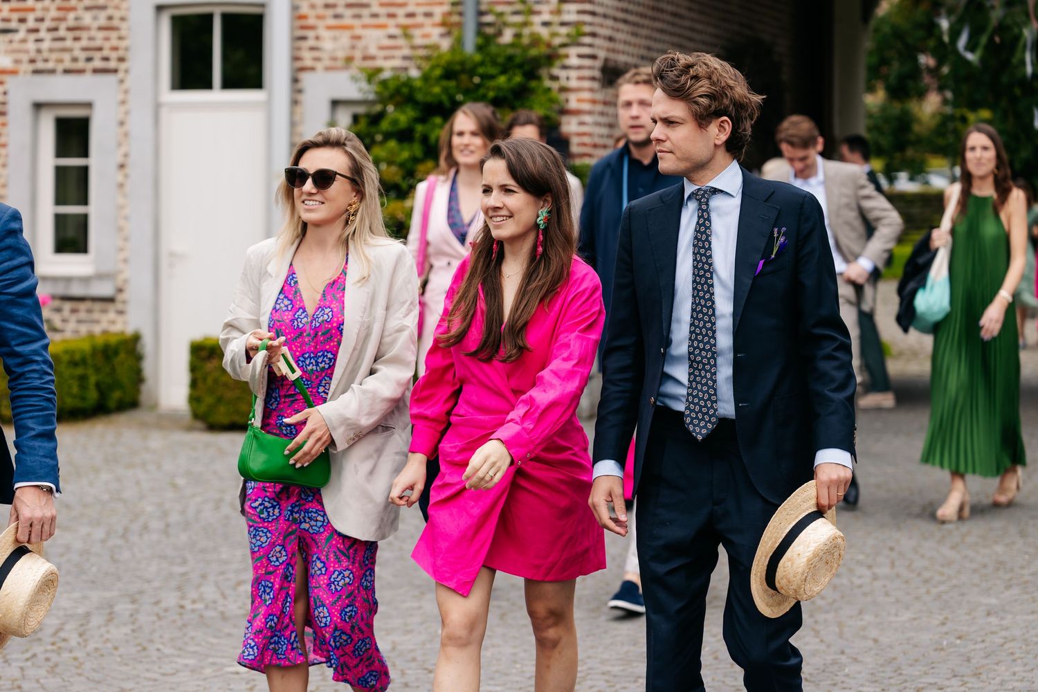 Elegantly dressed wedding guests walking across a cobblestone courtyard in colorful summer outfits.