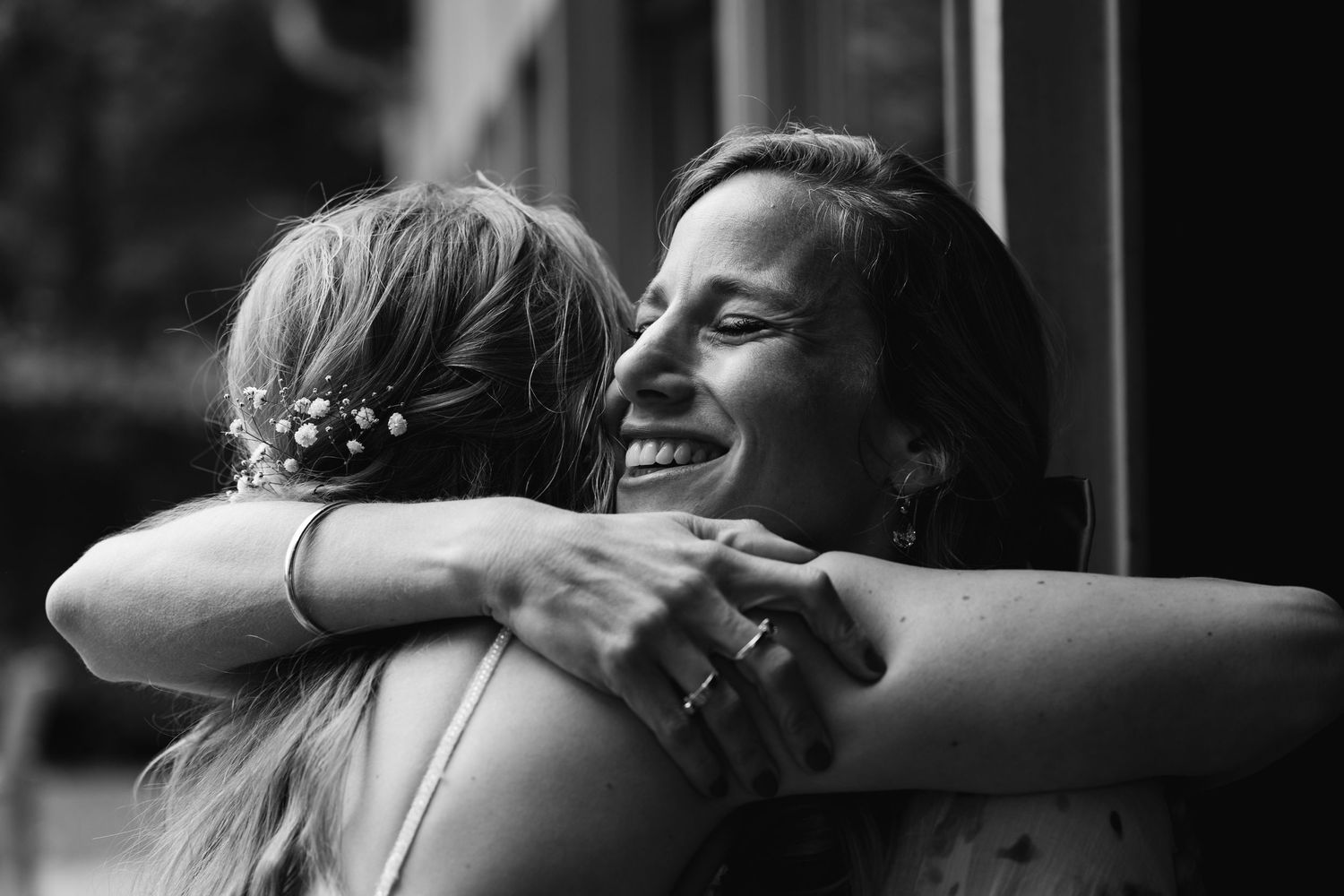 Two women share a warm embrace, smiling joyfully in a black and white wedding photo.