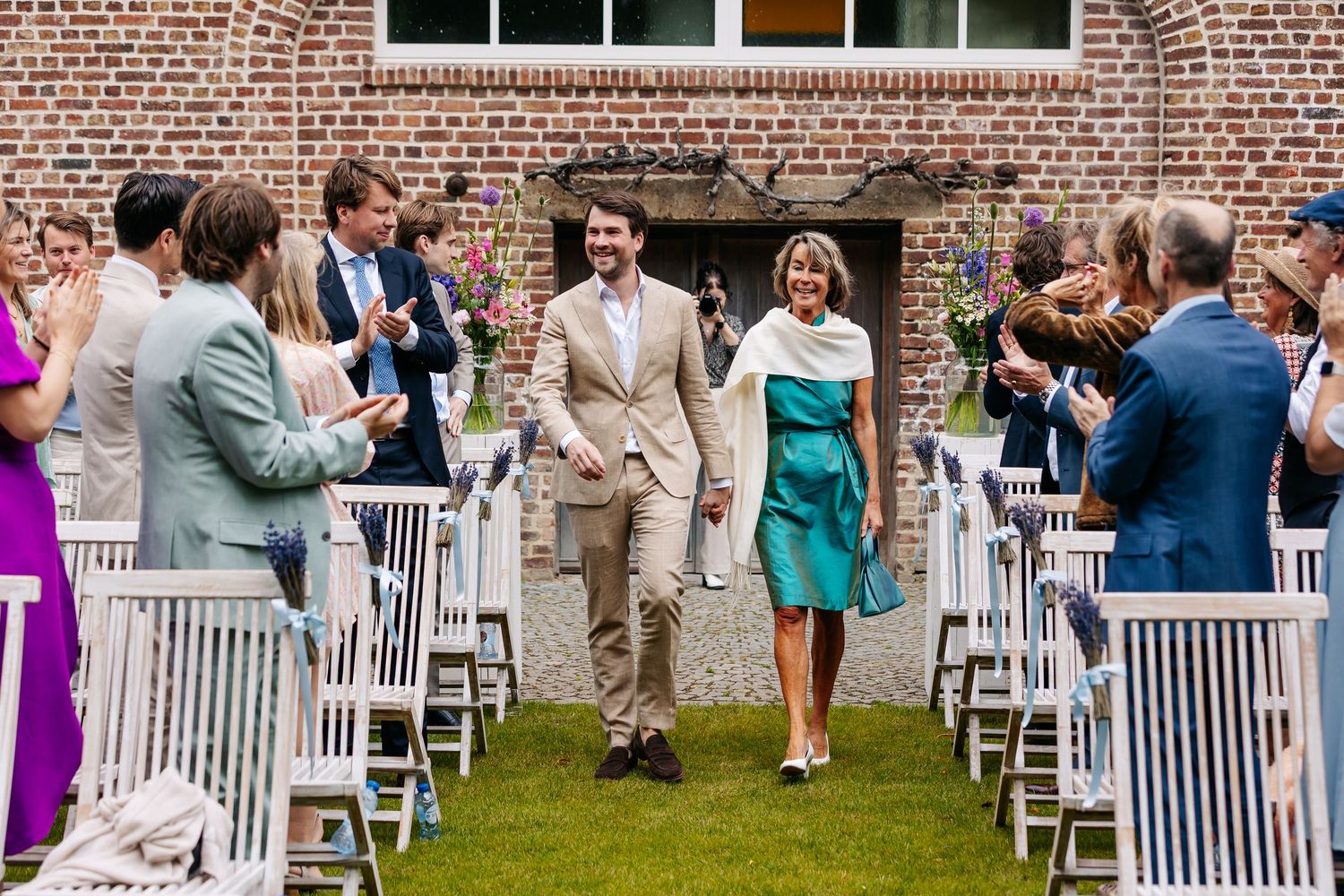 Smiling groom escorts elegantly dressed woman down outdoor wedding aisle as guests applaud.