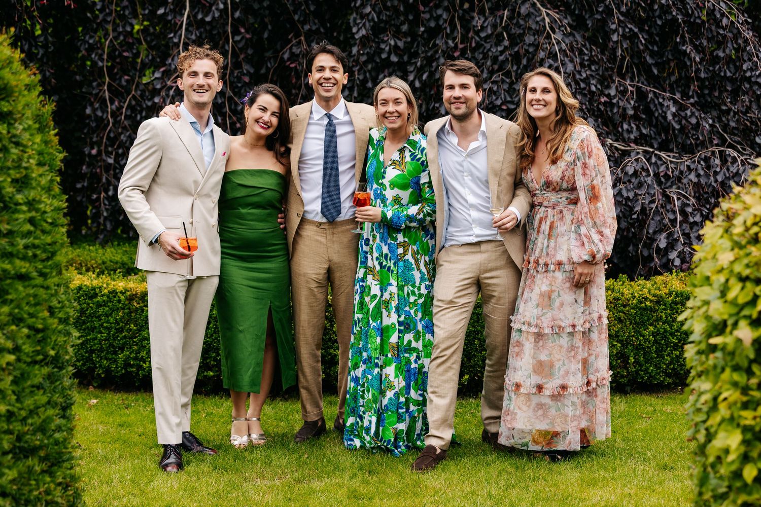 Six elegantly dressed wedding guests pose together smiling on a green lawn at an outdoor reception.