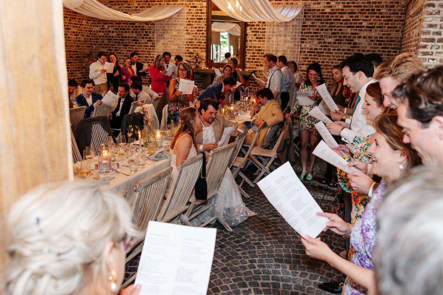 Wedding guests singing from song sheets during a candlelit indoor reception in a rustic brick venue.