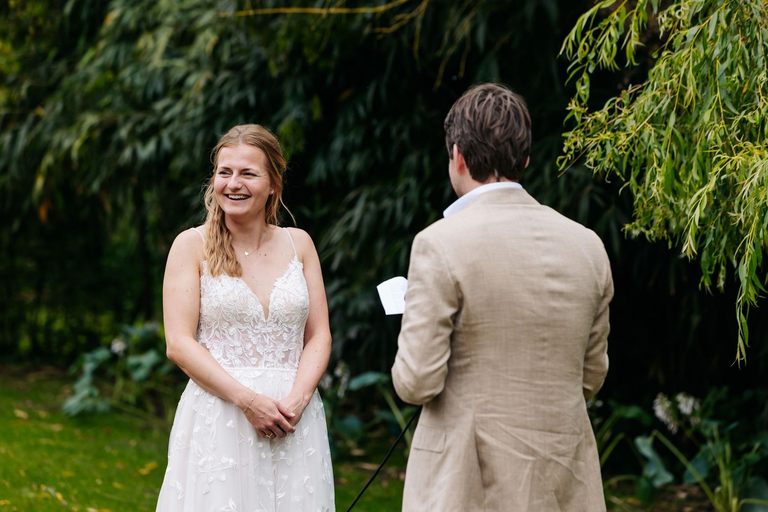 Bride in lace wedding dress smiling at groom reading vows in a lush garden setting.