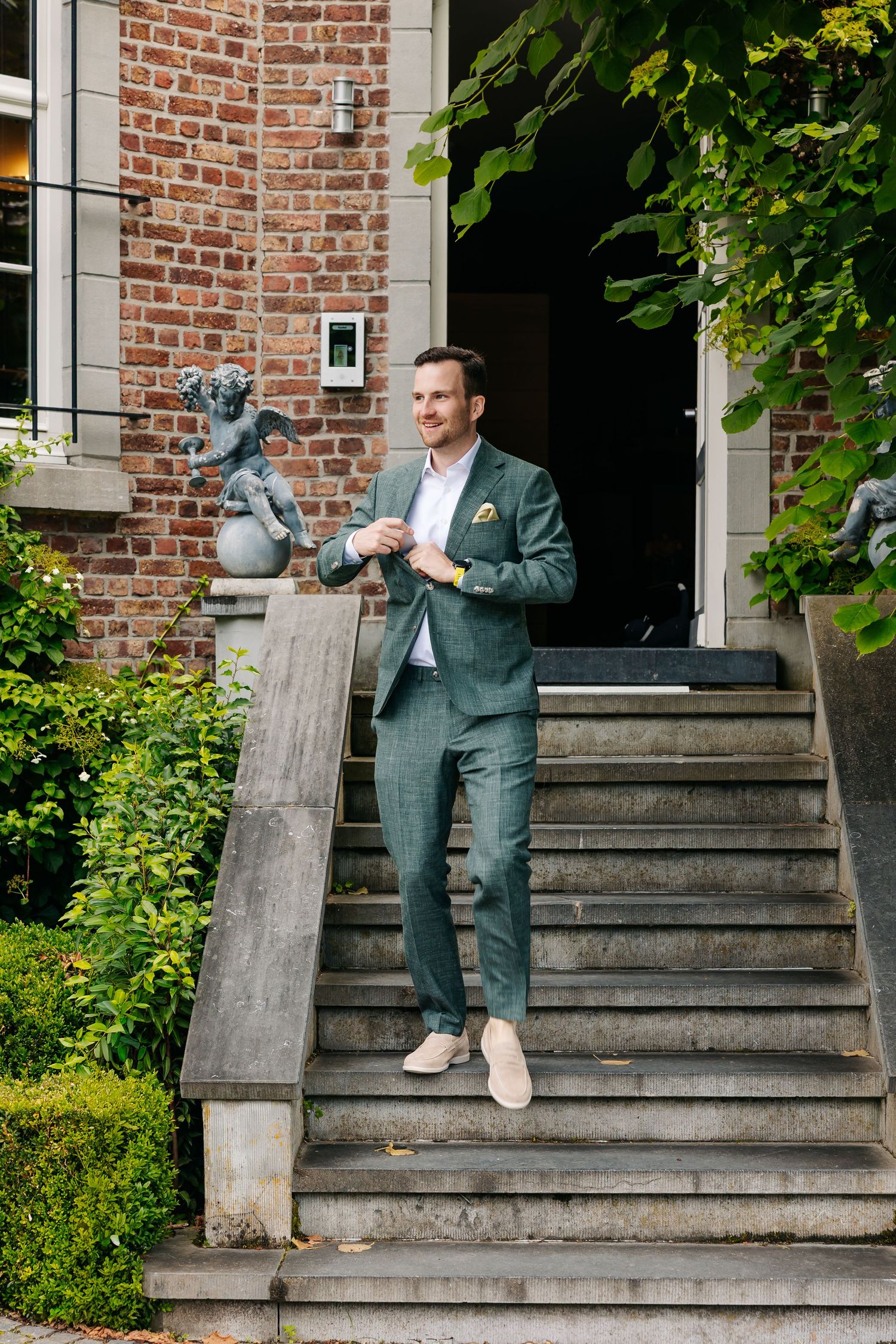 Stylish groom in a green suit descending stone steps outside a brick building with garden statues.