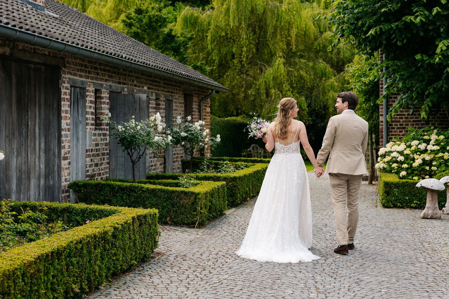 Bride and groom walk hand-in-hand through a charming cobblestone garden path.
