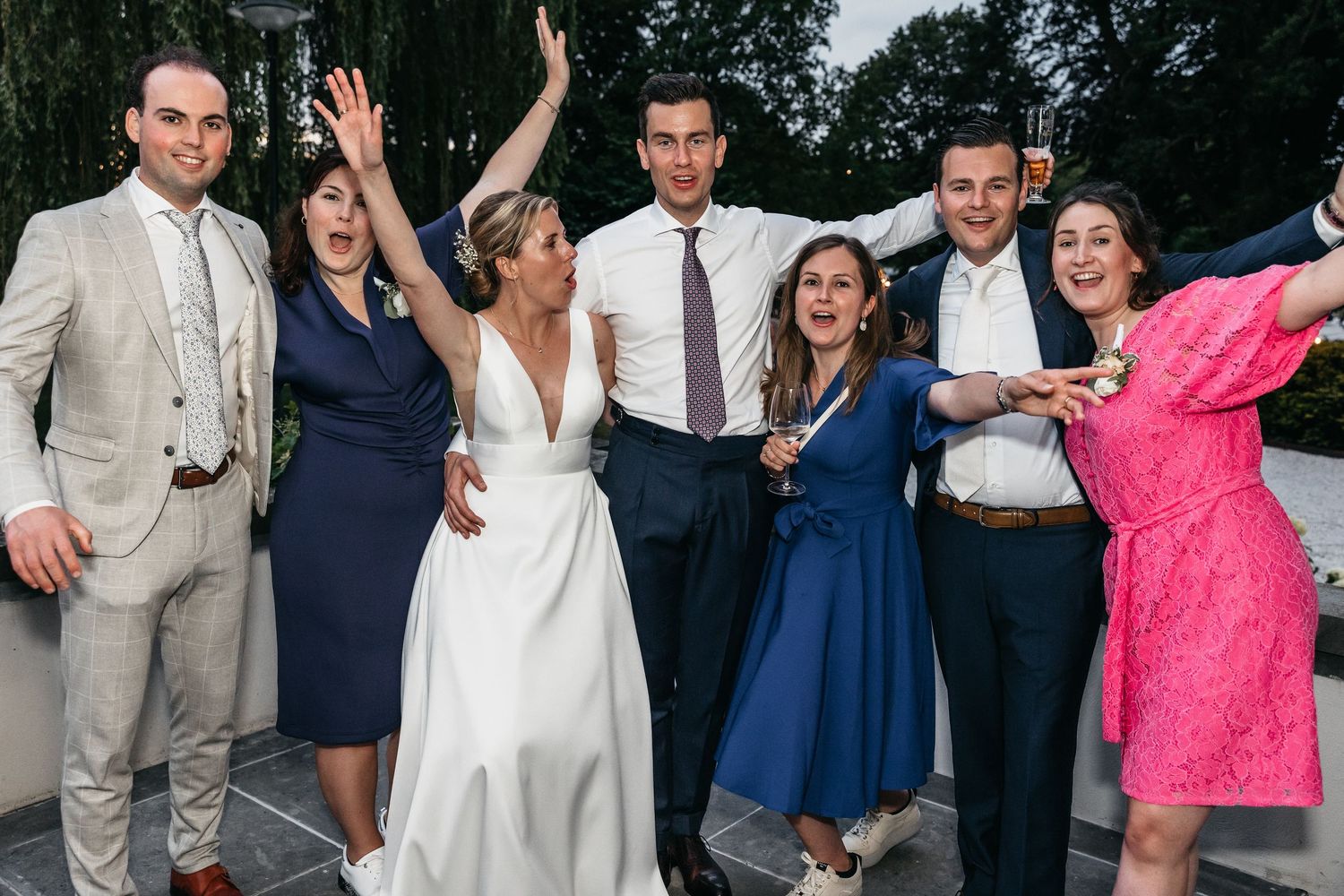 Bride and groom pose joyfully with wedding guests raising hands outdoors at evening reception.