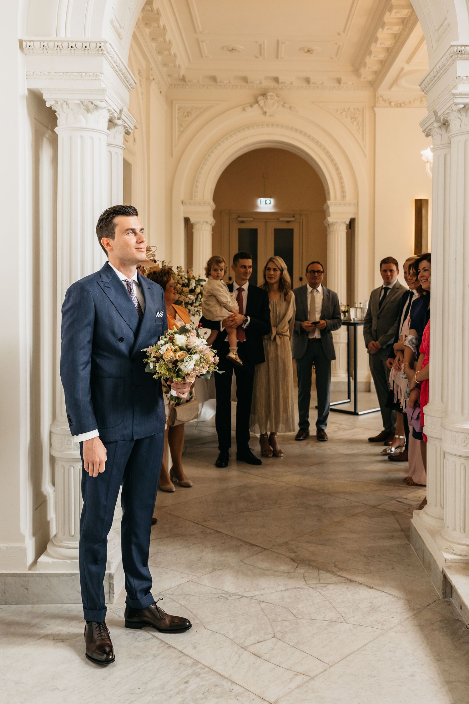 Groom in navy suit holding bridal bouquet awaiting bride in ornate hall.