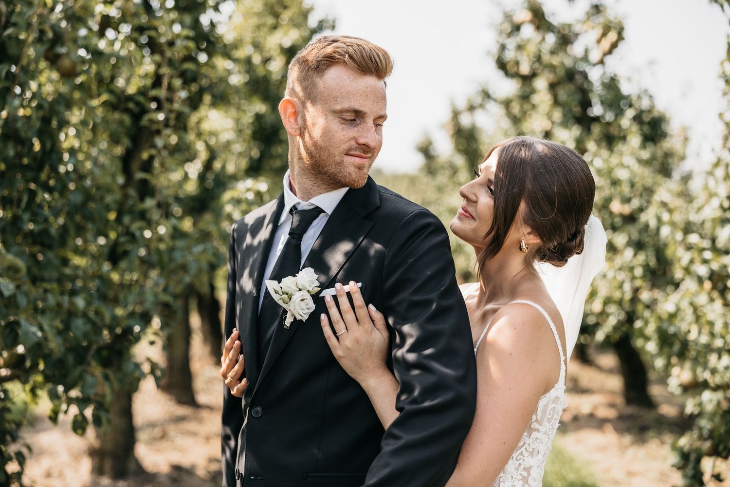 Bride laughs joyfully while standing close to groom with white boutonniere in sunny orchard.