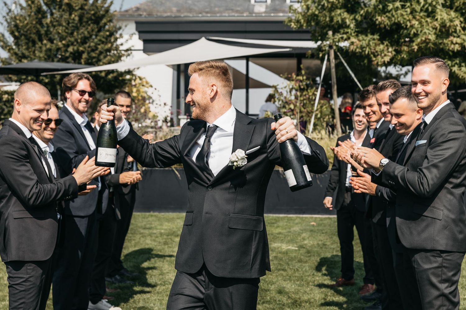 Groom opening champagne bottle surrounded by cheering groomsmen in black suits outdoors.