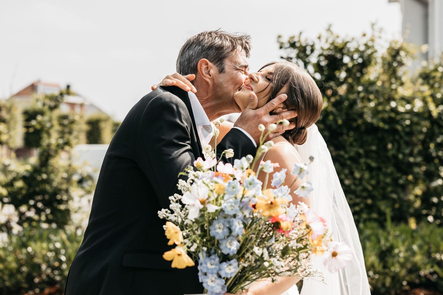 Bride and groom sharing romantic kiss while holding colorful wildflower bouquet in lush garden wedding setting.