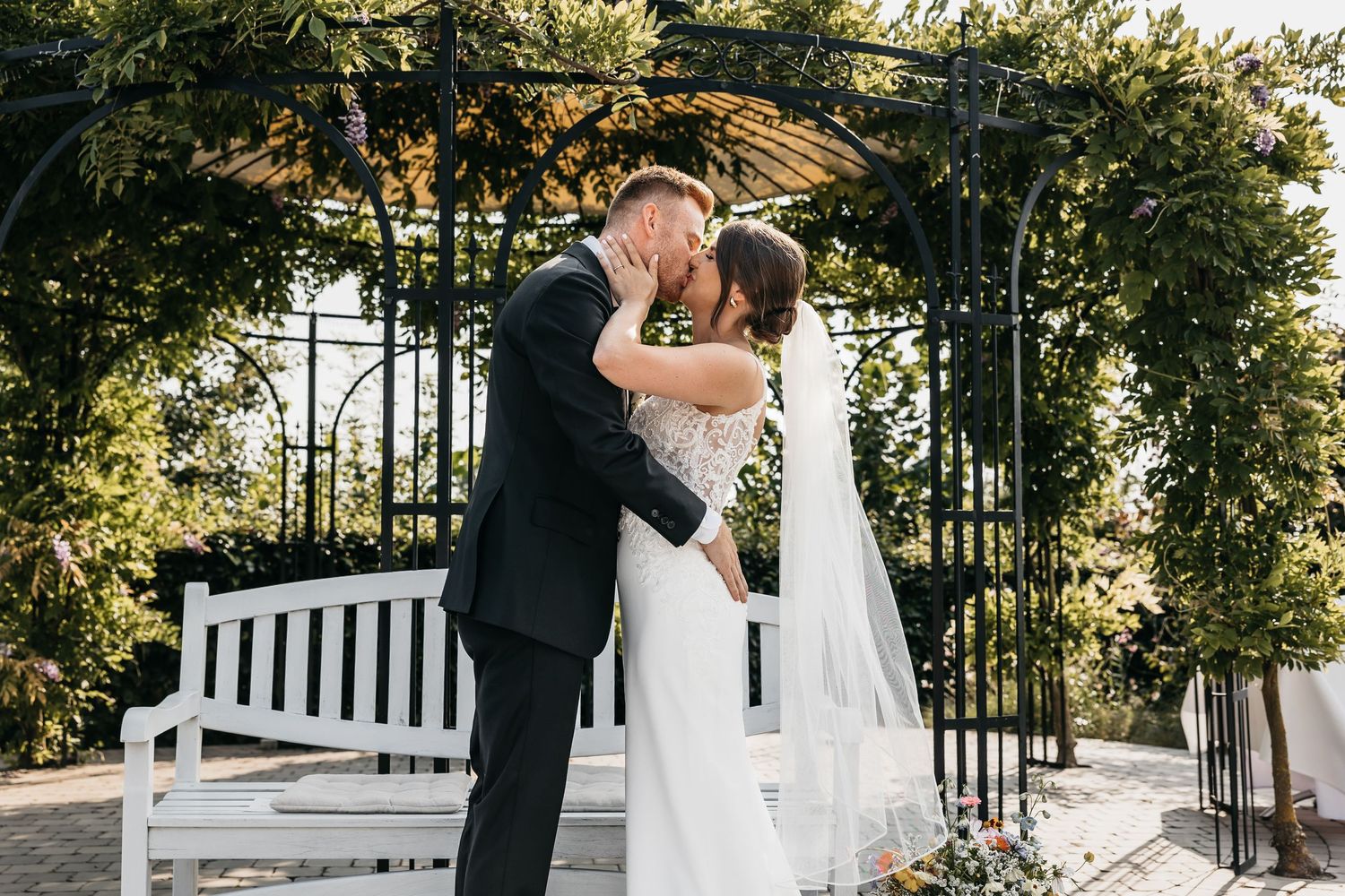Newlyweds share first kiss as husband and wife beneath romantic garden arbor outdoors.