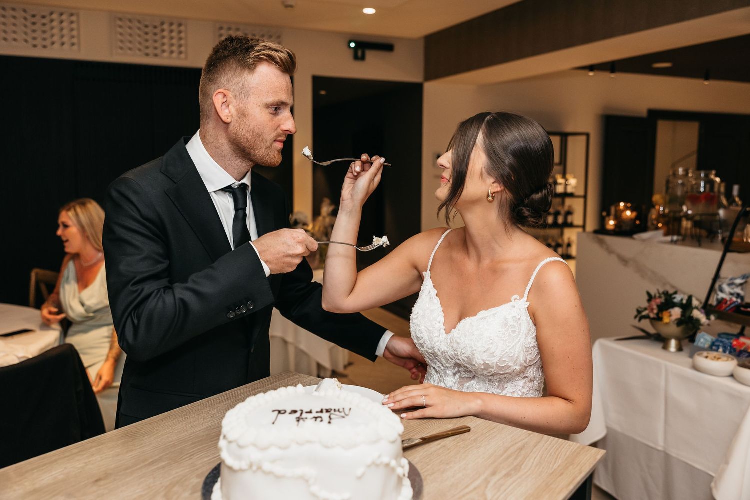 Groom feeding bride a bite of white wedding cake during indoor wedding reception celebration.