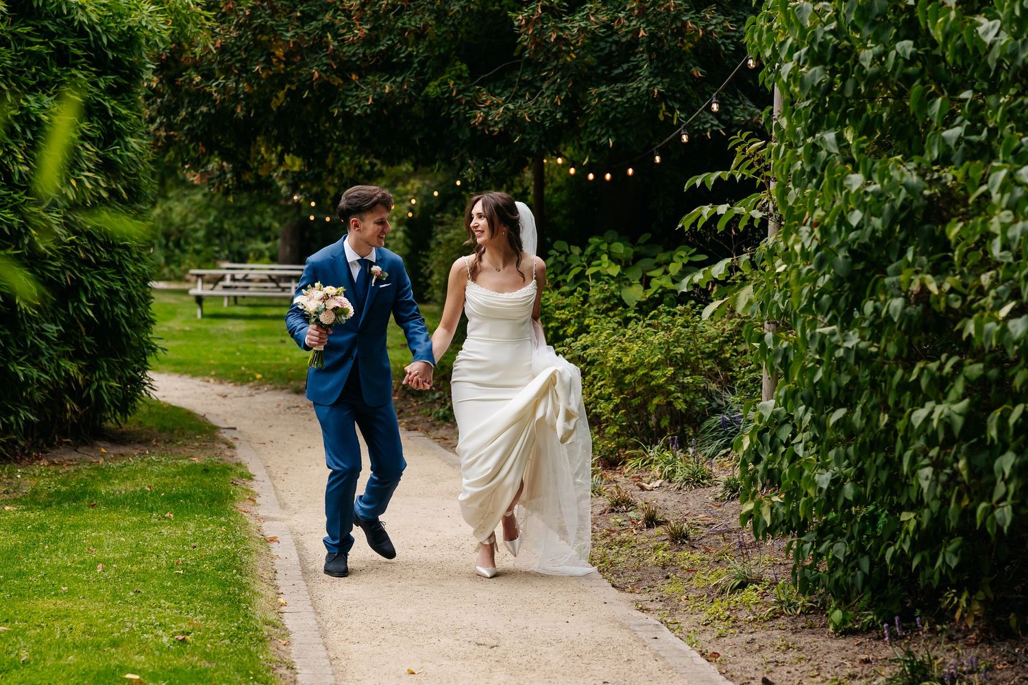 Happy bride and groom walking hand in hand along a garden path on their wedding day.