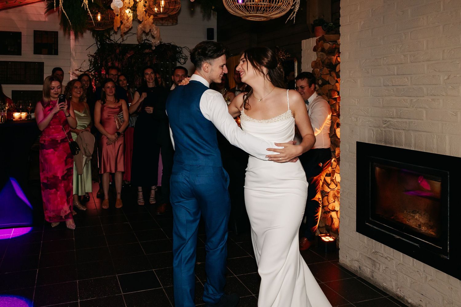 Bride and groom dance closely together near fireplace at wedding reception while guests watch and celebrate.