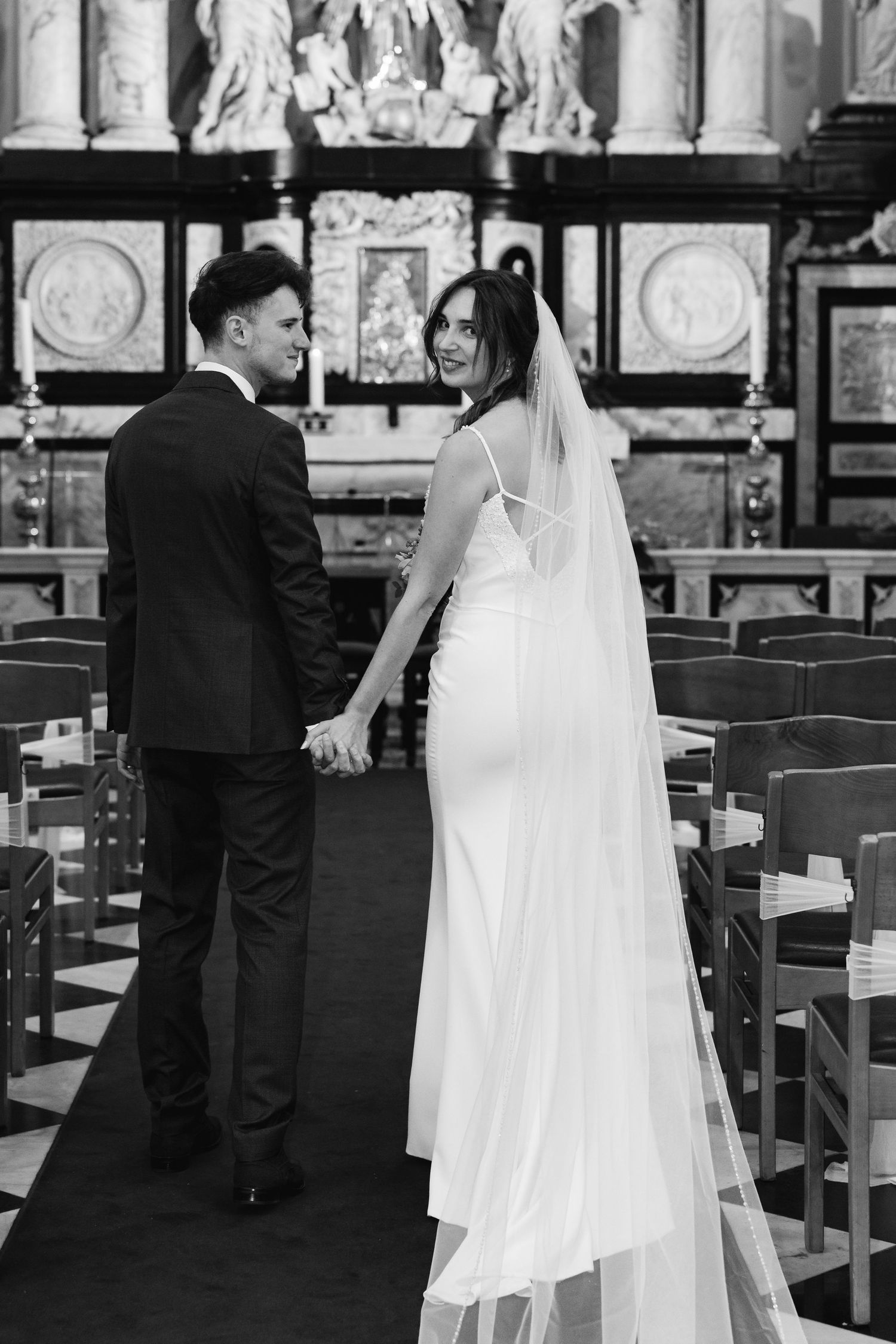 Black and white photo of newlyweds holding hands and smiling in elegant church.