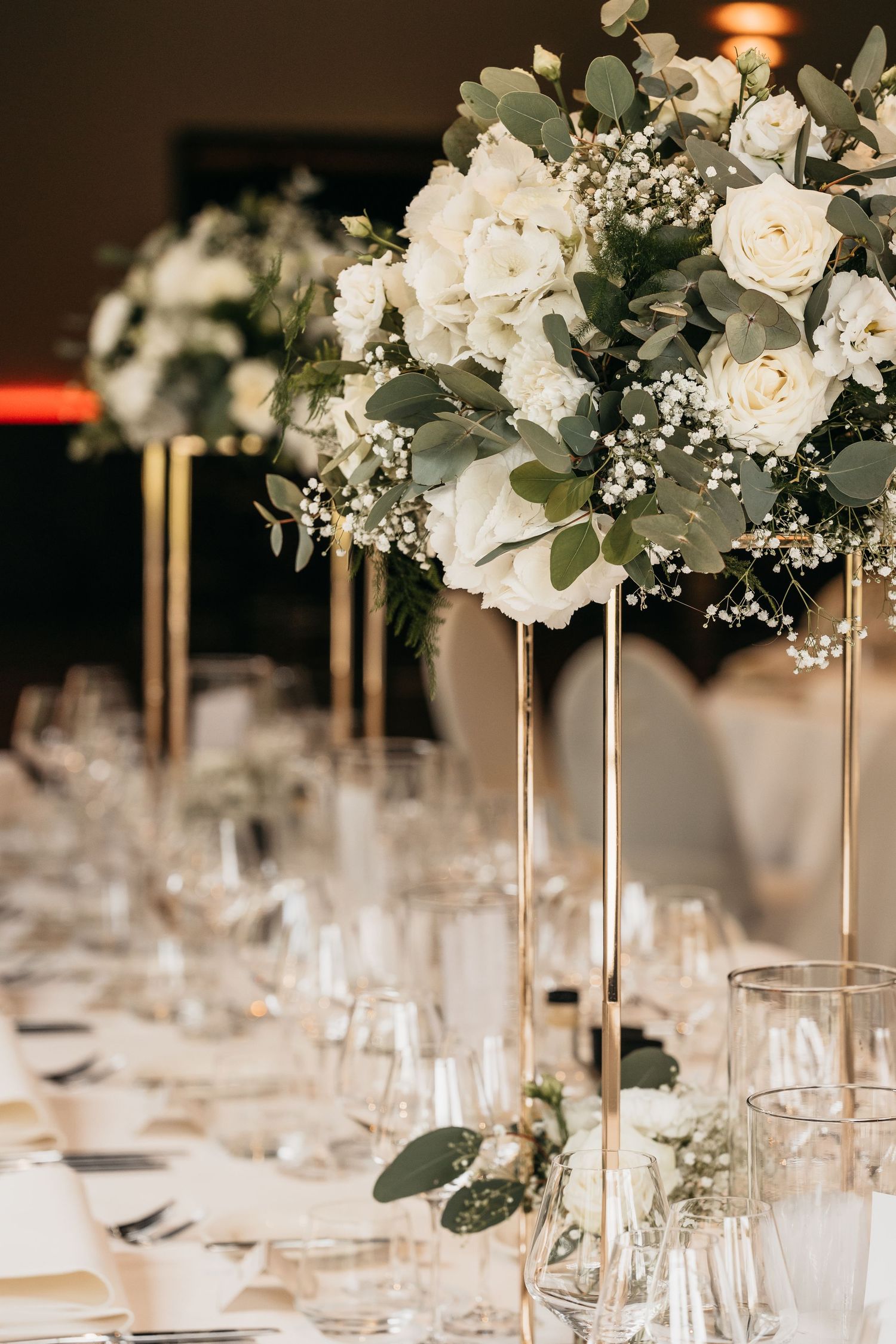Elevated gold floral centerpiece with white roses and eucalyptus above a elegantly set wedding reception table.