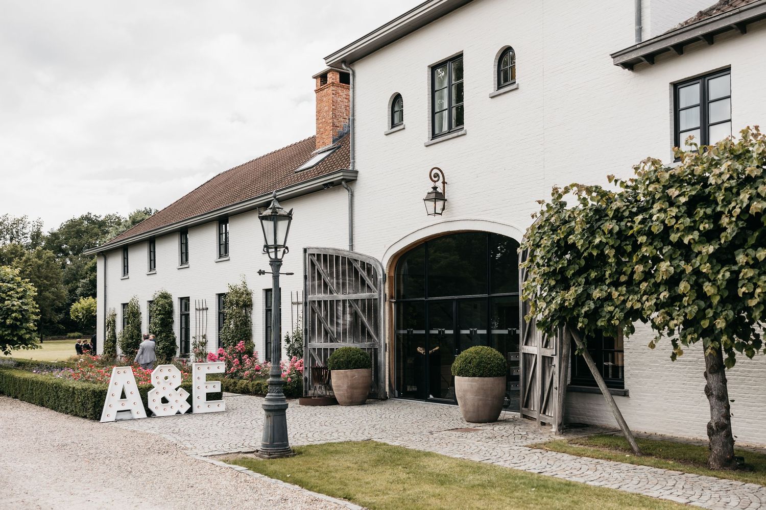 Elegant white farmhouse wedding venue with A&E marquee letters beside arched entrance gate.