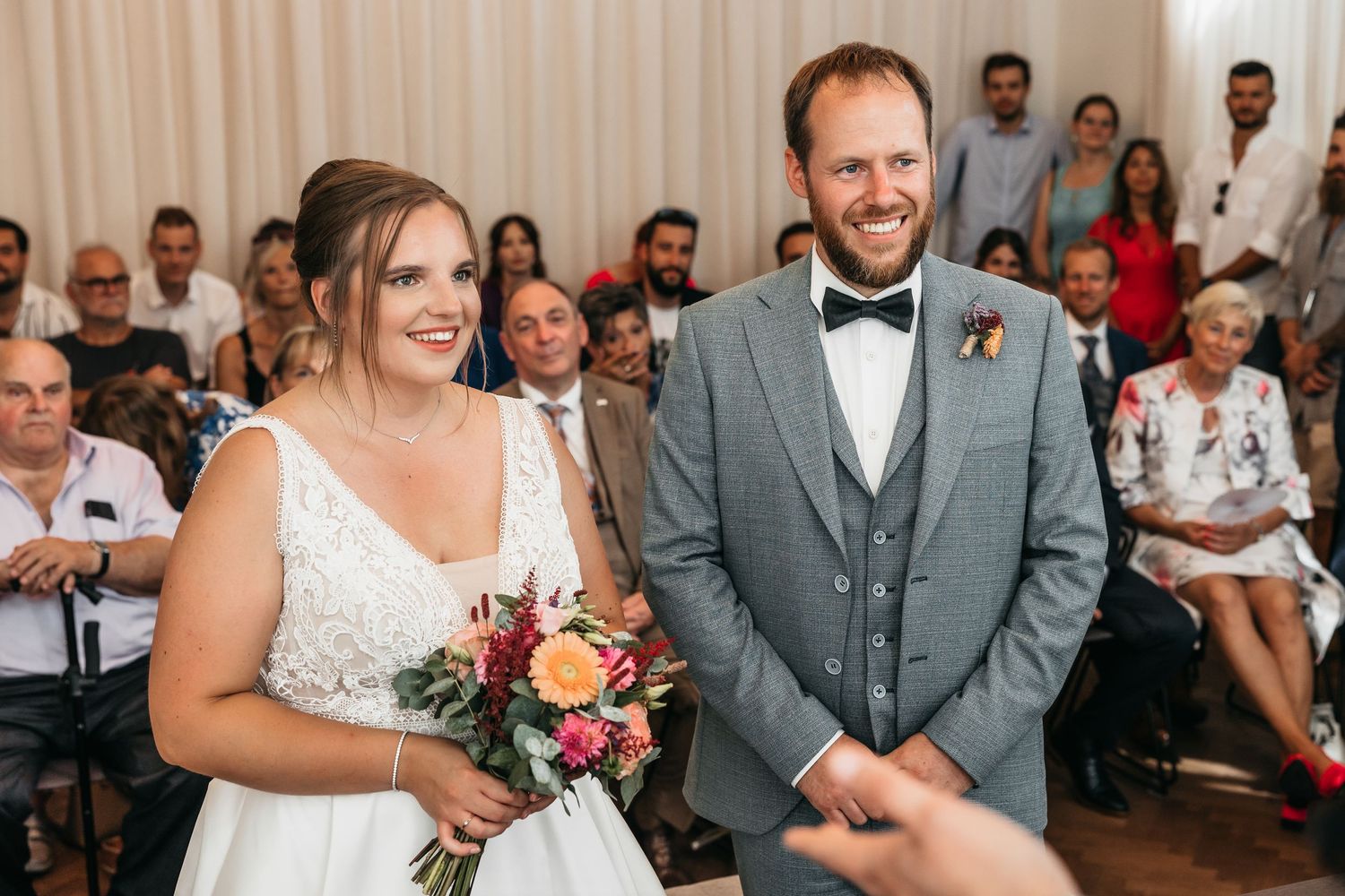 Smiling bride holding colorful bouquet stands beside groom in gray suit at wedding ceremony.
