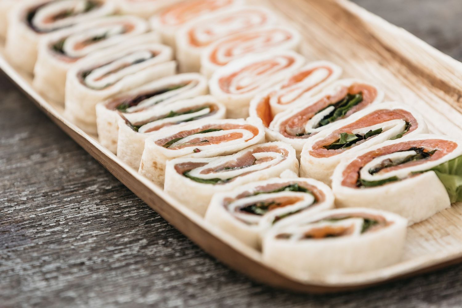 Assorted pinwheel wraps with smoked salmon and greens served on a wooden platter.