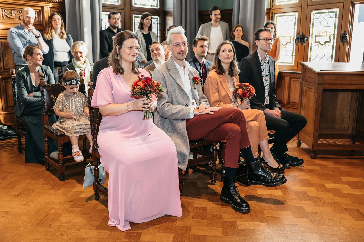 Wedding guests in pink and orange outfits seated with red bouquet at an elegant ceremony venue.